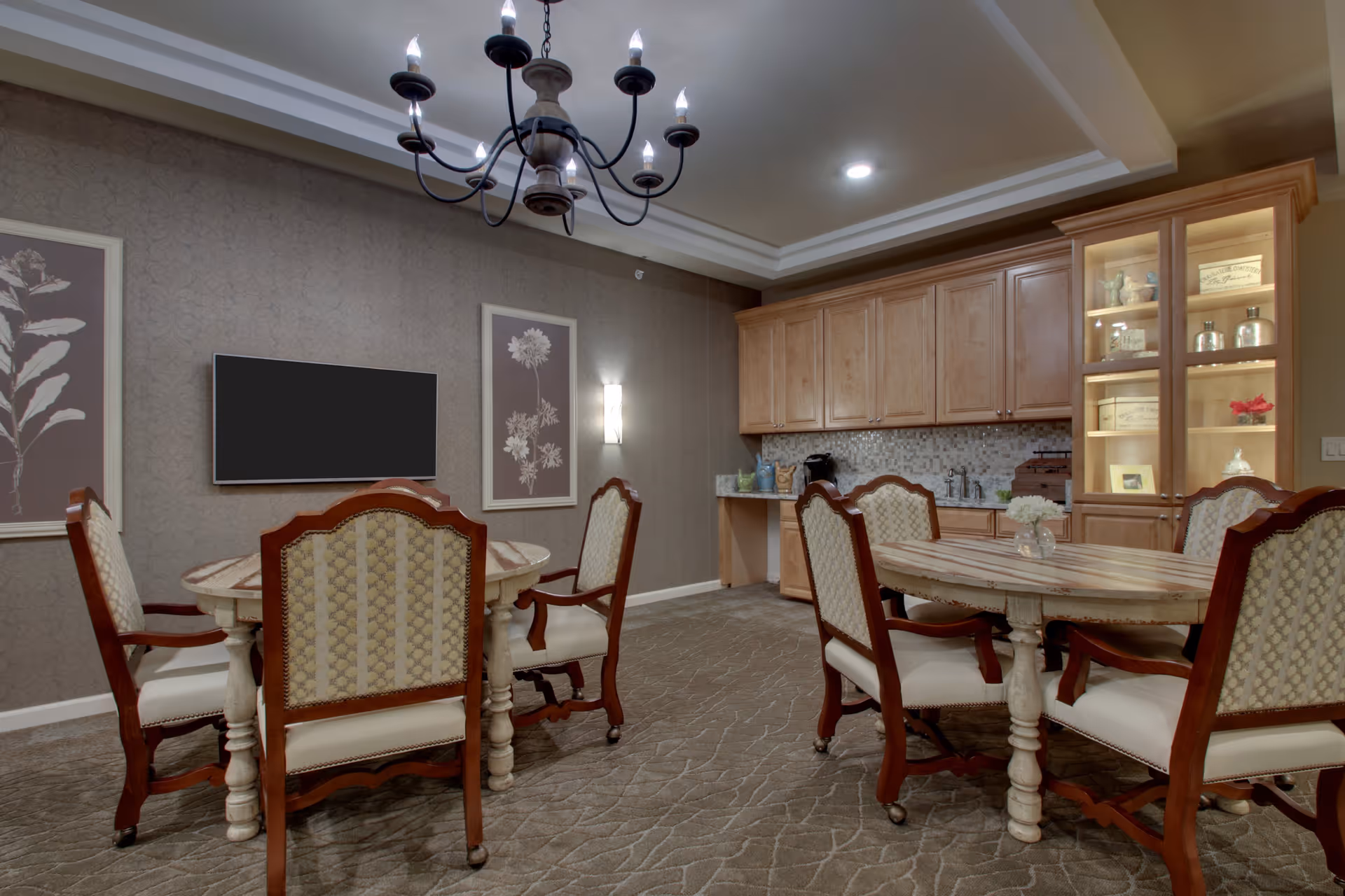 A cozy dining area in a senior living facility featuring two round wooden tables surrounded by upholstered chairs with wooden frames. The room has a patterned carpet, a chandelier hanging from the ceiling, framed botanical artwork on the walls, a flat-screen TV mounted on the wall, and a wooden cabinet with glass doors displaying decorative items. The back wall has a countertop with a mosaic tile backsplash and wooden cabinets above and below.