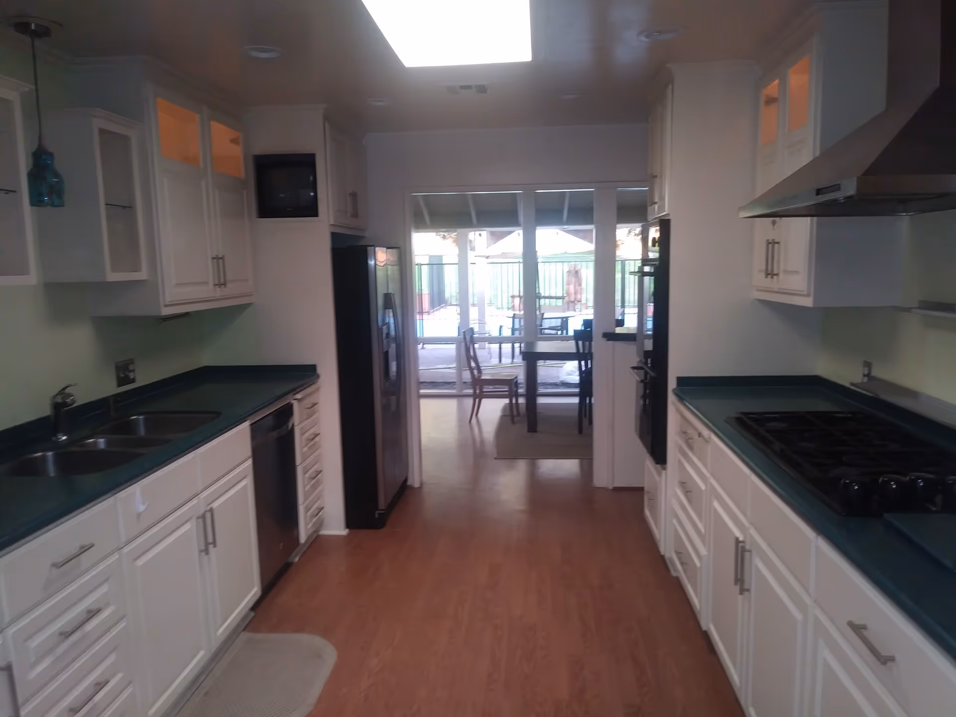 A kitchen with white cabinets and green countertops on both sides. There is a double sink and dishwasher on the left, and a stove with a range hood on the right. At the end of the kitchen, there is a dining area with a table and chairs, and large glass doors leading to an outdoor space with a pool and patio furniture.