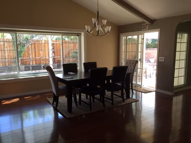 A dining room with a dark wooden table surrounded by six chairs, two of which have light upholstery. The room has wooden flooring and a large window with a view of a wooden fence outside. A chandelier hangs above the table, and there is a glass door leading to an outdoor patio area.