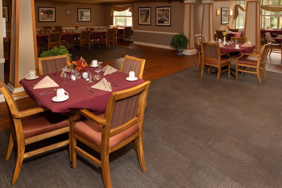 A dining room with several tables covered in burgundy tablecloths, each set with cups, glasses, napkins, and silverware. Wooden chairs with cushioned seats surround the tables. The room features carpeted and hardwood flooring, framed artwork on the walls, and large windows letting in natural light.