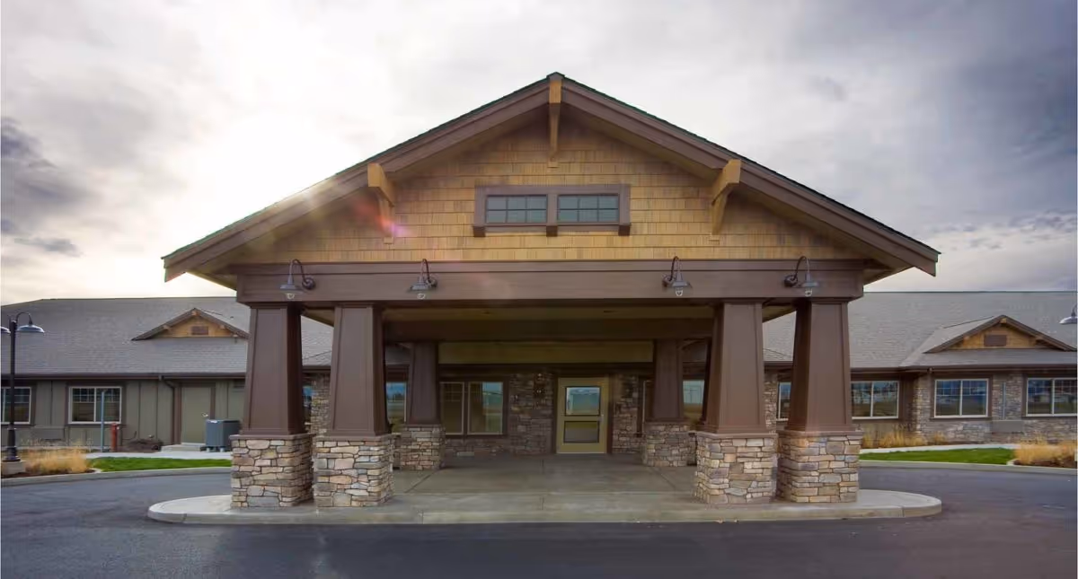 Front exterior view of Creekside Inn Memory Care Community building with a covered entrance supported by stone and wood pillars under a cloudy sky.