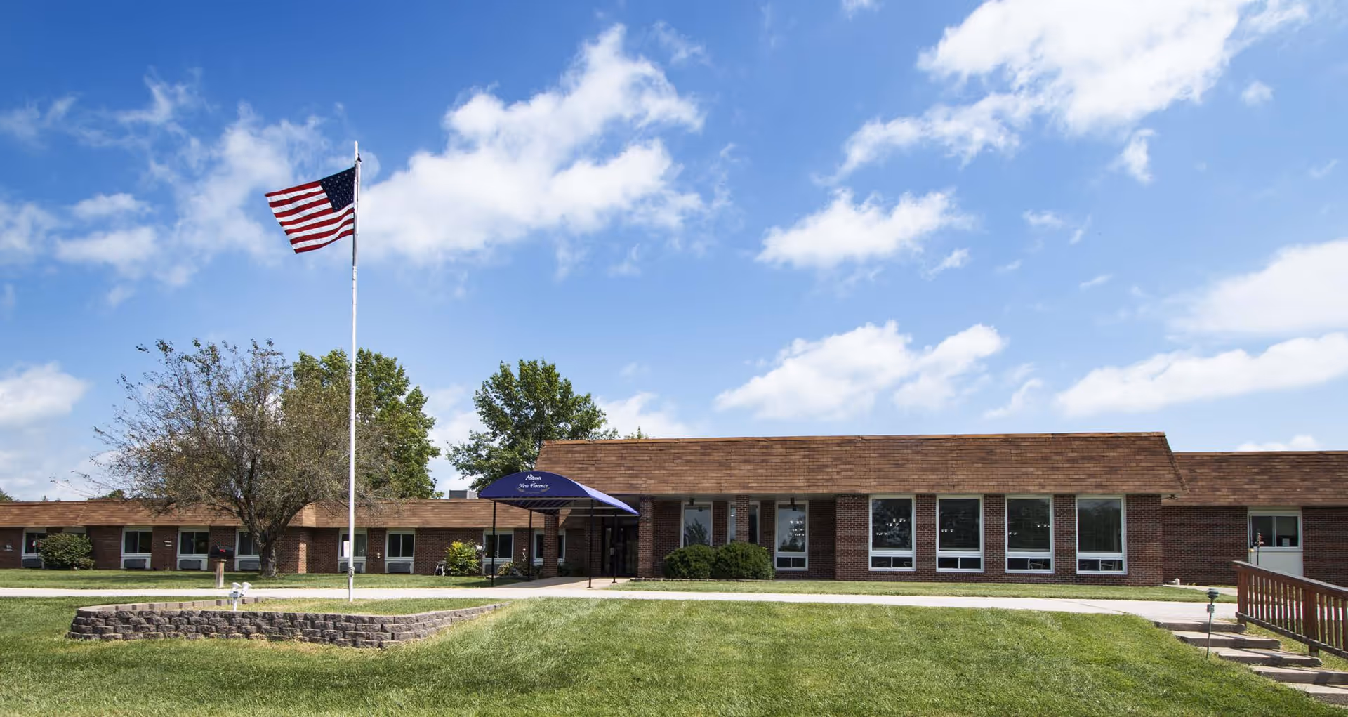 Exterior view of St. Andrew's at New Florence facility with a brick building, a blue awning over the entrance, an American flag on a flagpole, green lawn, and a partly cloudy blue sky.
