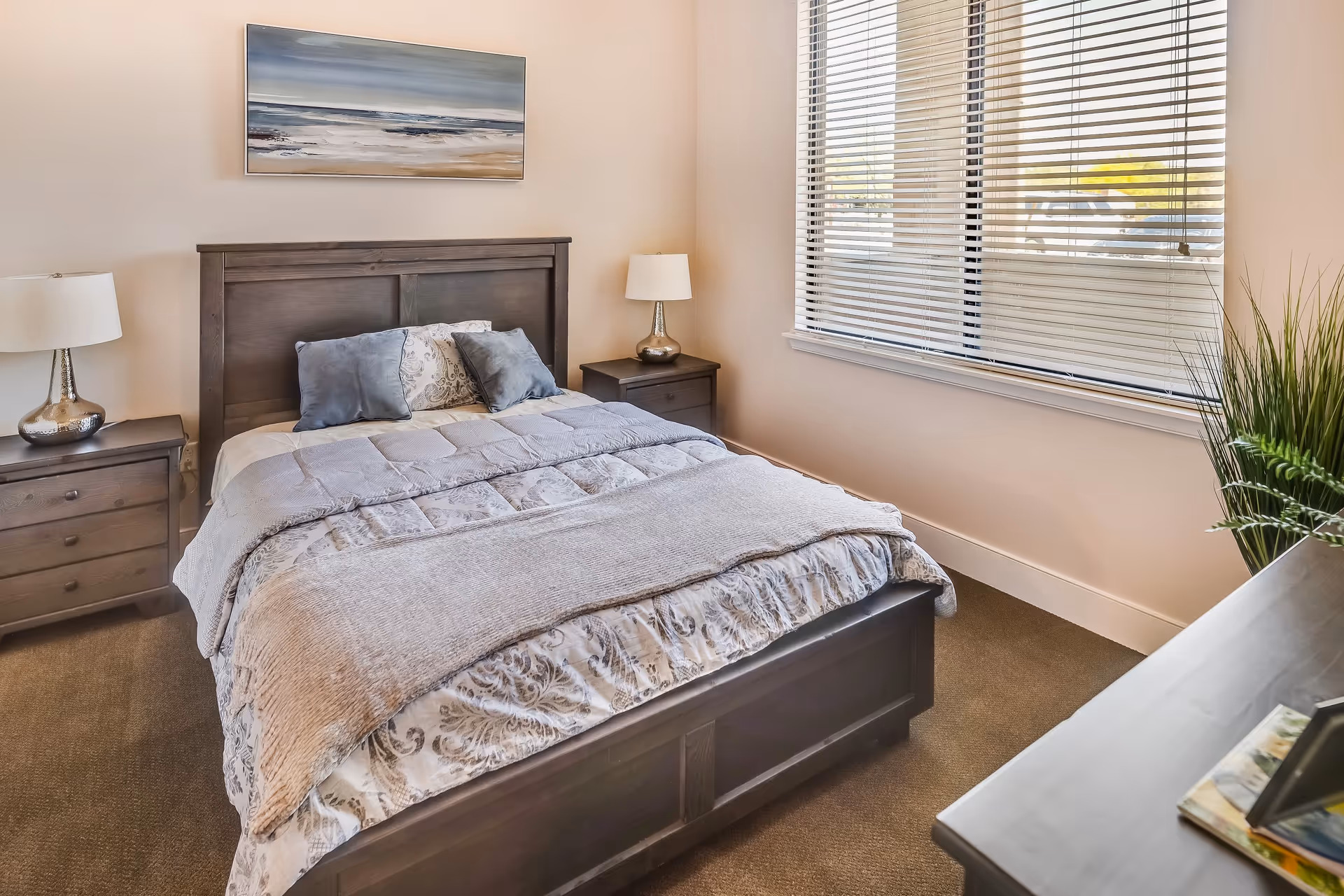 A neatly made bed with a wooden headboard and patterned bedding in a bedroom. There are two bedside tables with lamps on each side of the bed. A large window with blinds is on the right wall, and a framed painting of a beach scene hangs above the bed. A plant and some books are on a dresser in the foreground.