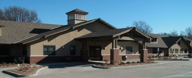 Exterior view of a single-story building with a covered entrance, beige walls with brick accents, and a small tower-like structure on the roof under a clear blue sky.