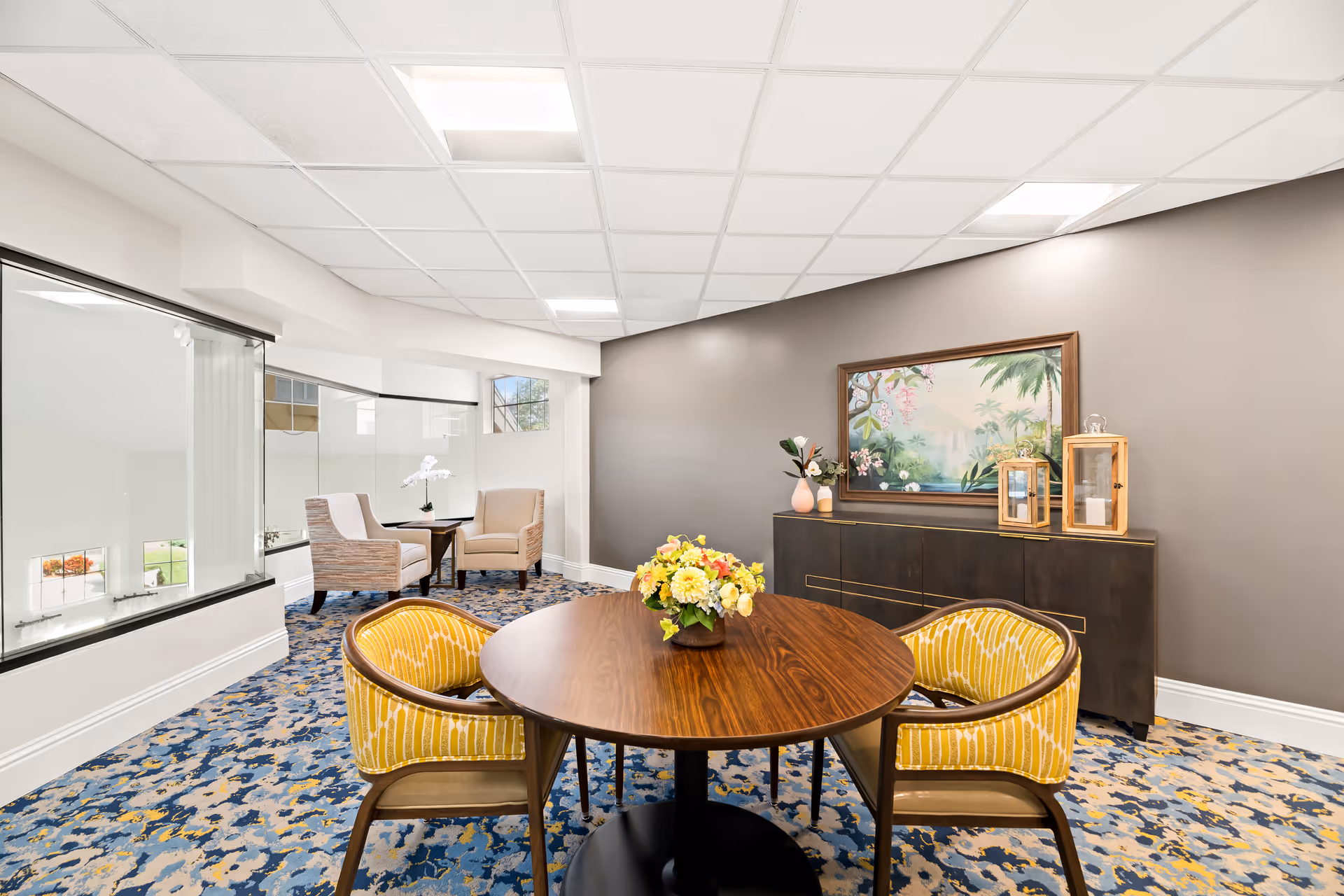 A bright and cozy interior room with a round wooden table surrounded by four yellow cushioned chairs. In the background, there are two beige armchairs with a small table between them near large windows. A dark wooden sideboard against the wall holds decorative items including vases and lanterns, with a framed tropical-themed painting hanging above it. The floor is covered with a patterned carpet in shades of blue and beige, and the ceiling has white tiles with recessed lighting.
