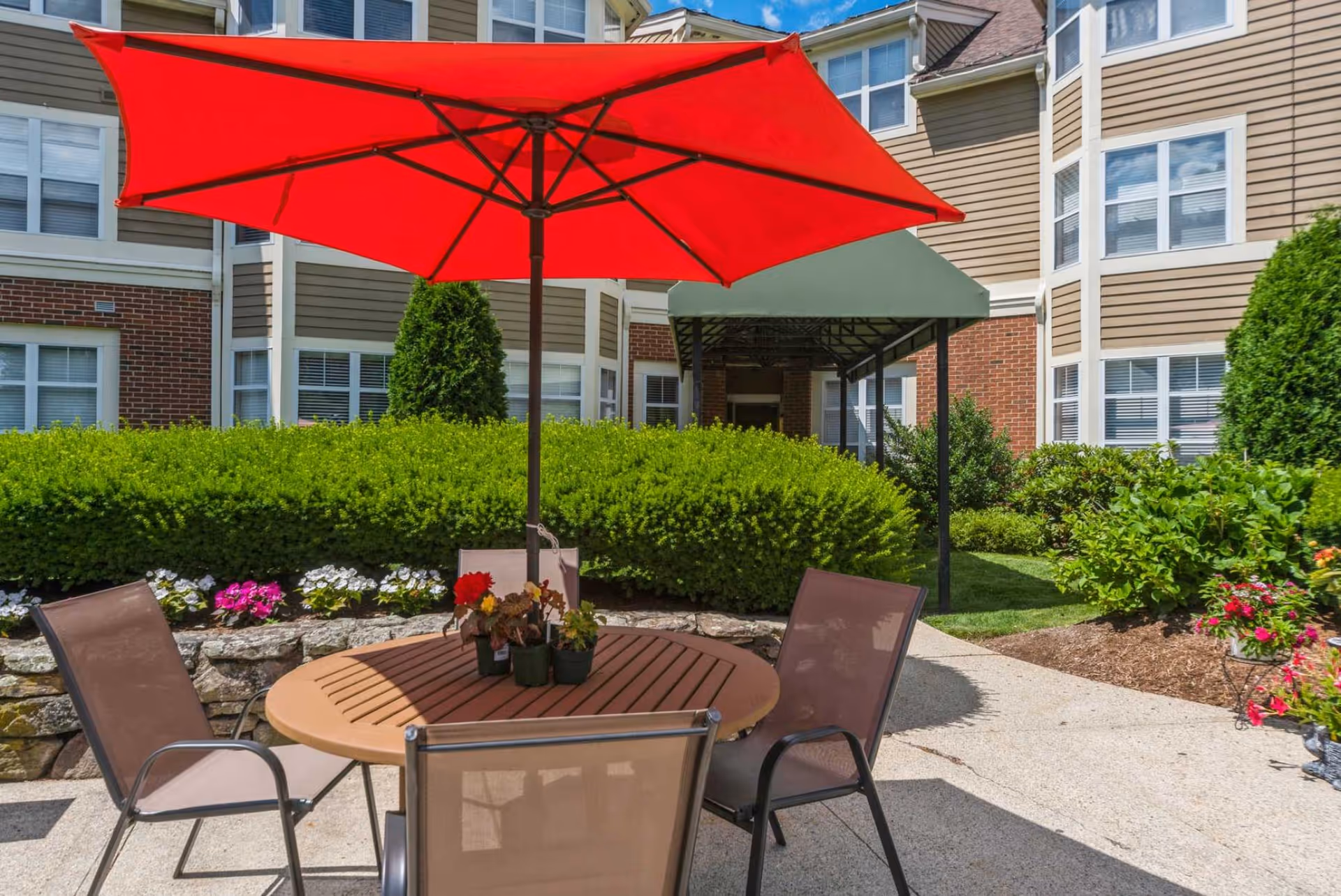 Outdoor patio area at The Gables at Winchester featuring a round wooden table with four chairs and a large red umbrella. The patio is surrounded by well-maintained bushes, flowers, and the exterior of the building with multiple windows.