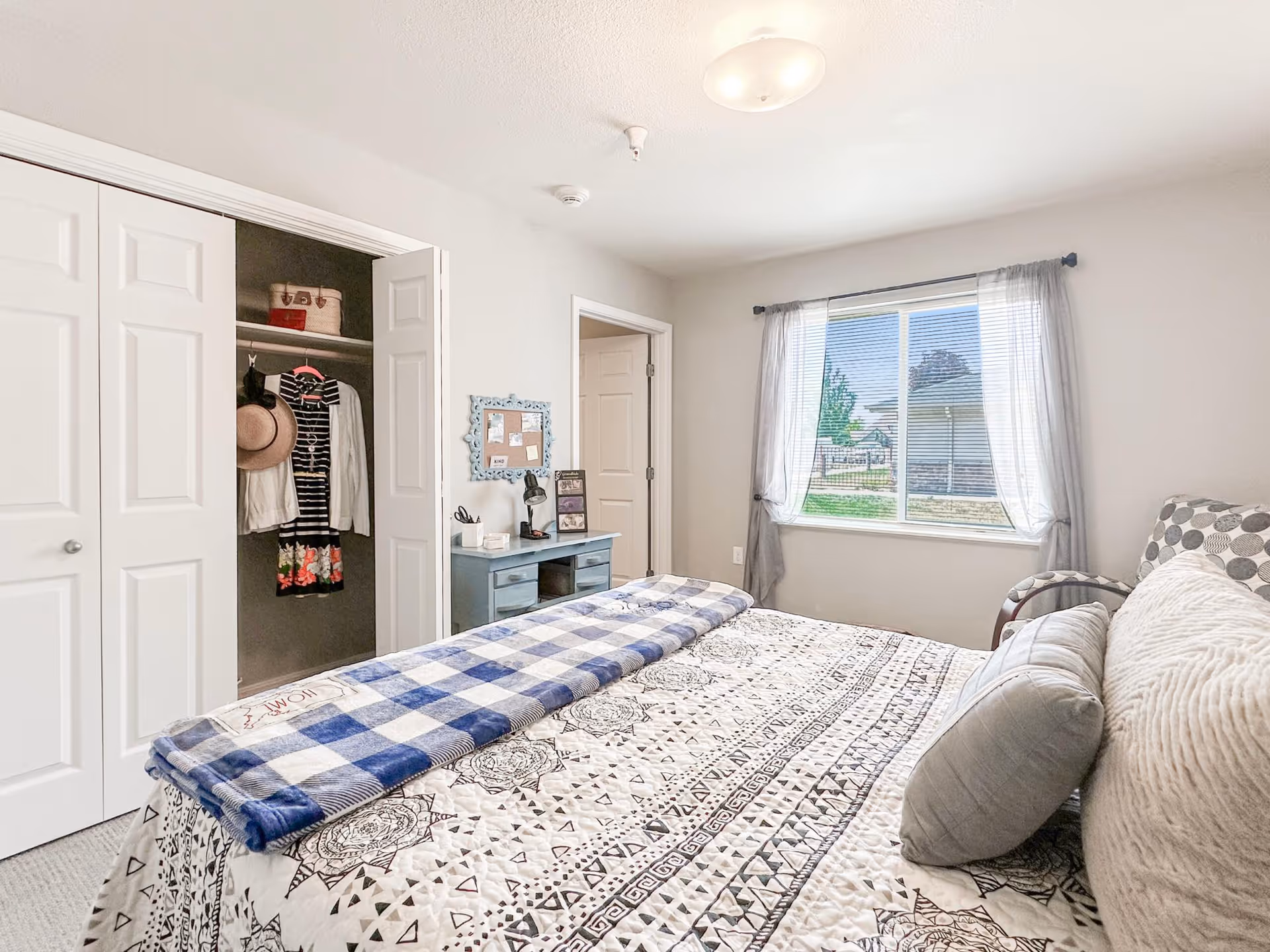 A bright bedroom with a patterned bedspread and multiple pillows on the bed. There is a blue checkered blanket folded at the foot of the bed. To the left, there is an open closet with clothes and a hat hanging inside. A small blue desk with a lamp, picture frames, and a bulletin board above it is positioned against the wall. A window with sheer curtains lets in natural light, showing a view of greenery and a neighboring building outside.