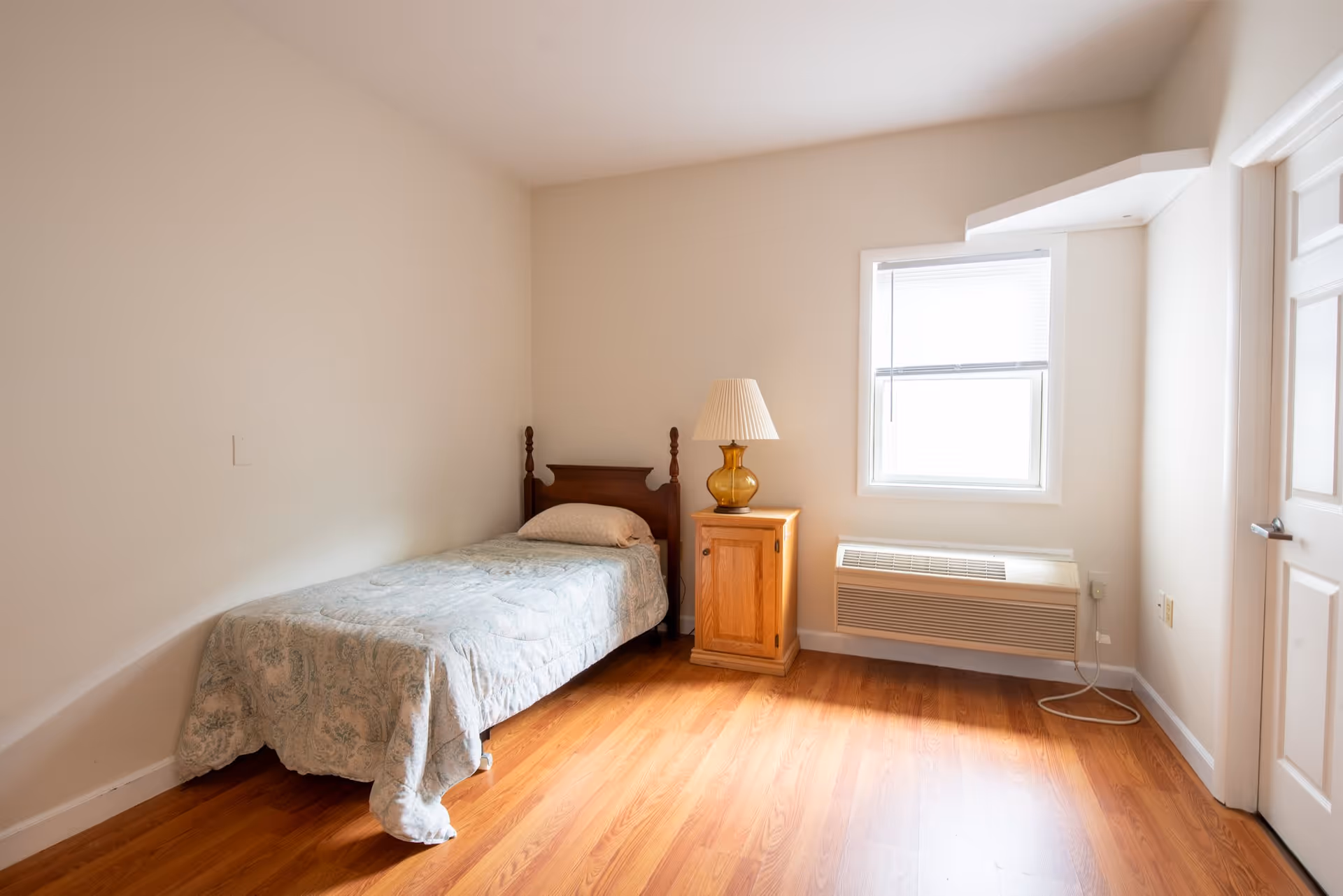 A simple bedroom with a single bed covered with a light blue patterned bedspread, a wooden nightstand with a yellow glass lamp, a window with blinds, and a wall-mounted air conditioning unit below the window. The room has light-colored walls and wooden flooring.