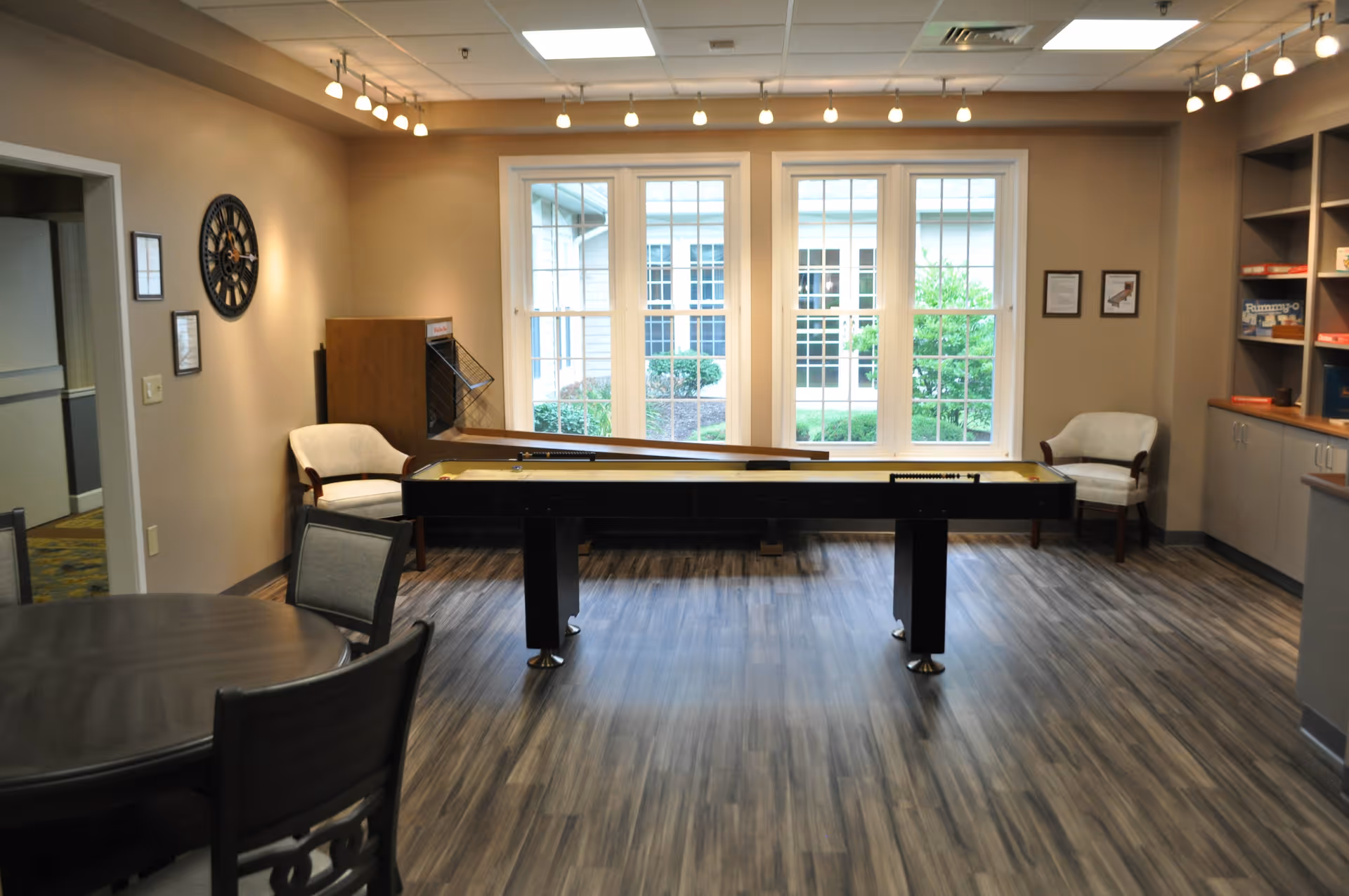 A well-lit recreational room with a shuffleboard table in the center, two armchairs placed near large windows showing an outdoor garden, a round table with chairs in the foreground, and shelves with board games on the right side.