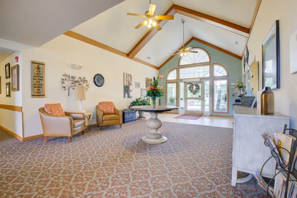 A spacious and well-lit lobby area with a high vaulted ceiling featuring wooden beams and ceiling fans. The room has patterned carpet flooring and light-colored walls adorned with decorative items including a clock, a metal tree wall art, and the word 'HOME'. There are two leather armchairs with orange patterned cushions, a round table with a flower arrangement in the center, and a white sideboard with decorative items on the right. Large windows and glass doors at the far end allow natural light to enter, showing an outdoor view.