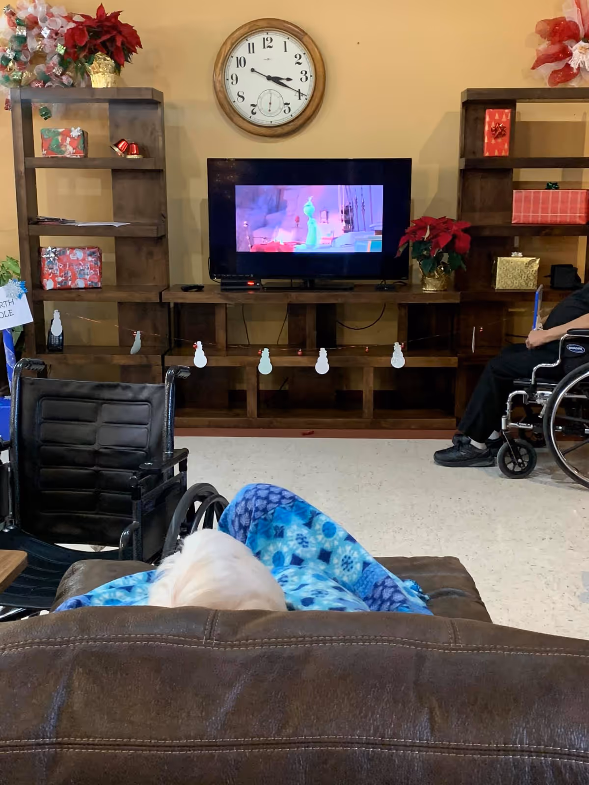 A cozy living room area in a senior living facility with a television mounted on a wooden entertainment center. The TV is playing an animated movie. There are holiday decorations including wrapped presents, poinsettia plants, and snowman garlands. Two wheelchairs are visible, one occupied by a person and the other empty. A person covered with a blue patterned blanket is seated on a brown leather couch facing the TV. A large round clock is mounted on the wall above the TV.