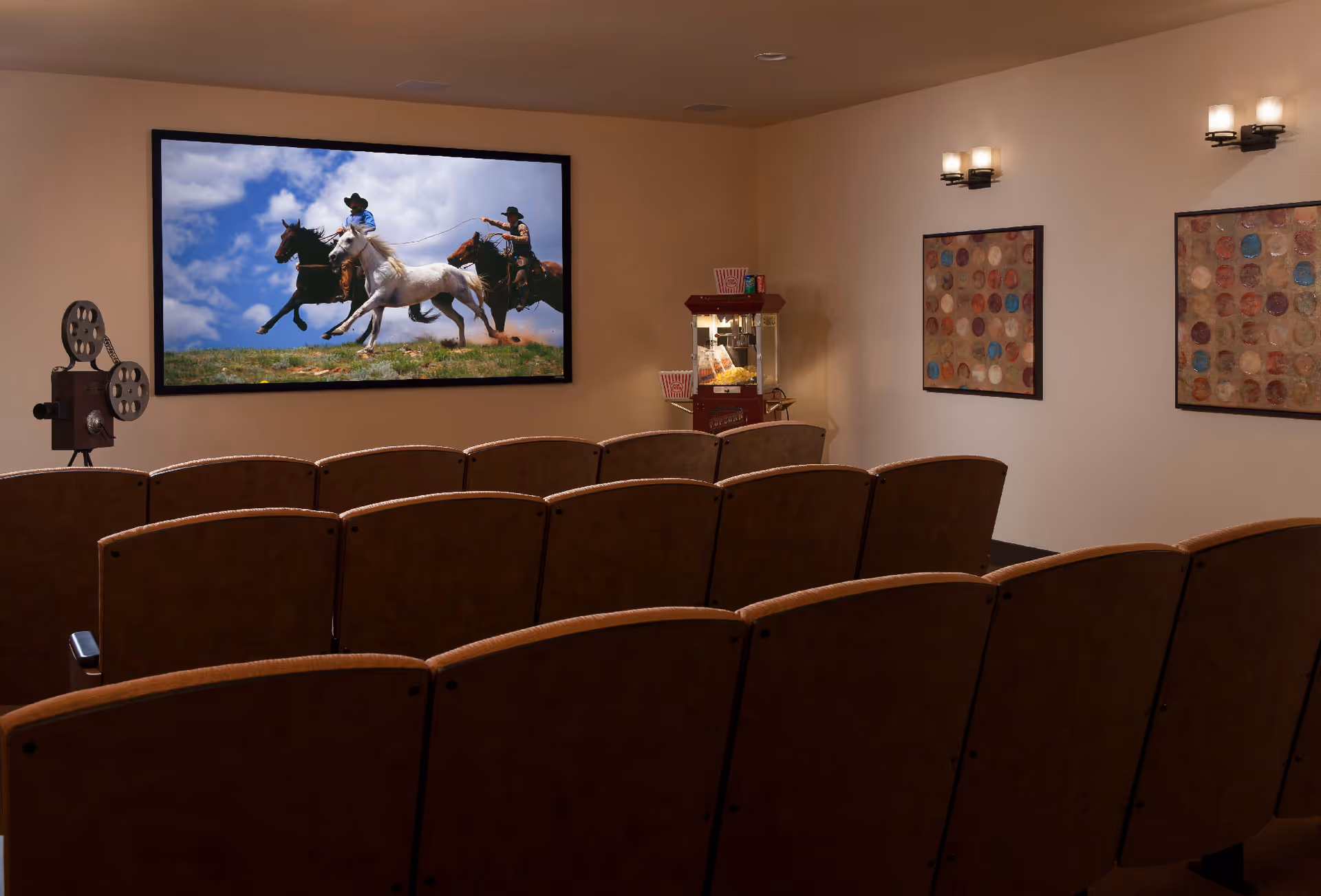 Small movie theater room with several rows of brown cushioned seats facing a large screen showing an image of three cowboys riding horses. There is a vintage film projector on the left side and a popcorn machine in the back corner. The walls are beige with two colorful abstract paintings and wall-mounted lights.
