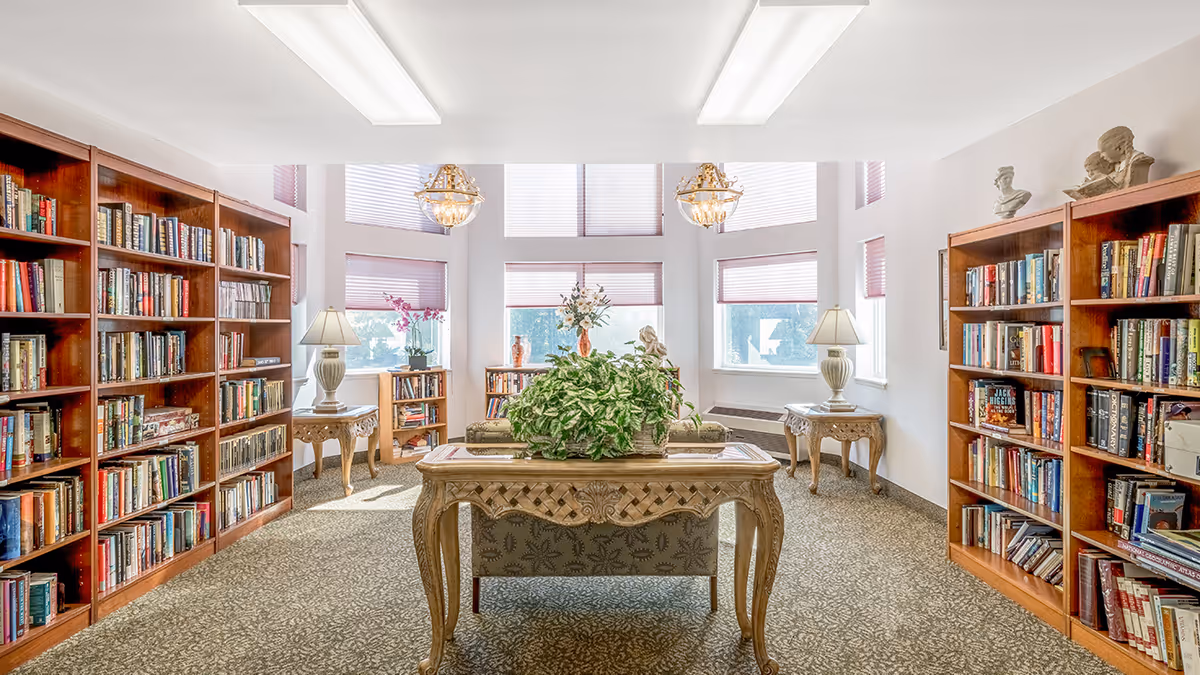 Sunlit communal library/reading room with bookshelves lining both sides, a decorative table with a plant in the center, lamps and seating by large windows.