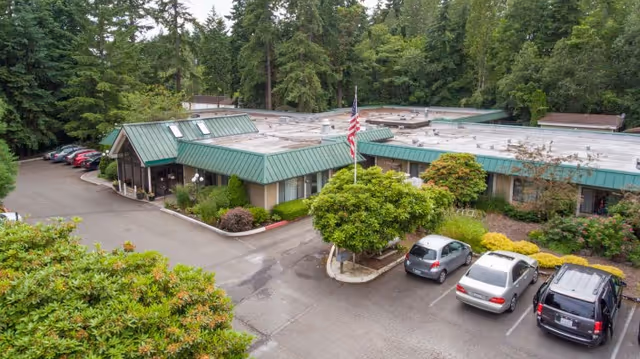 Exterior view of Bainbridge Island Health & Rehab Center showing a single-story building with a green metal roof, surrounded by trees and landscaping. There is a parking lot with several cars and an American flag on a flagpole near the entrance.