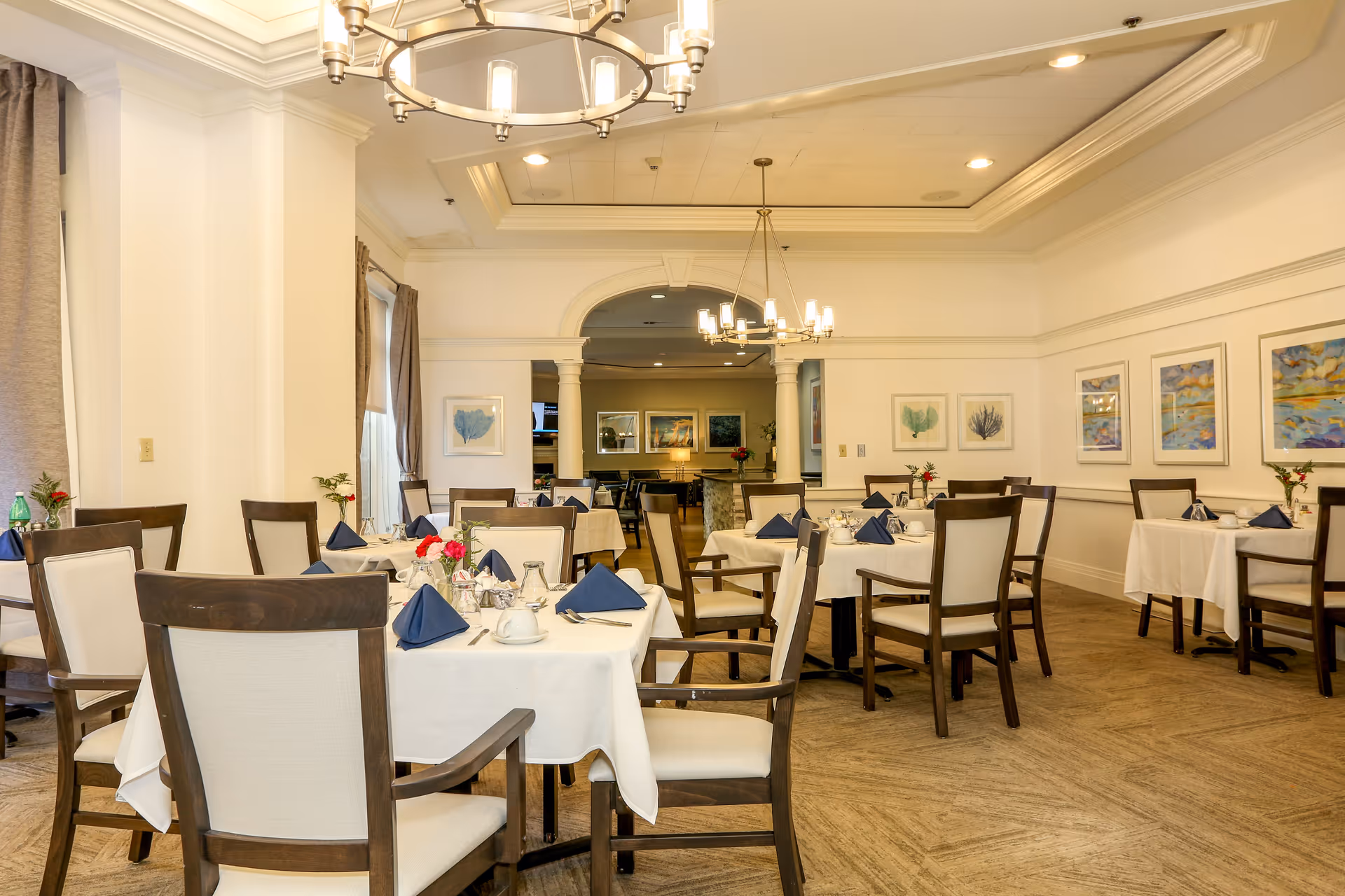 A bright and elegant dining room with multiple tables covered in white tablecloths, each set with blue folded napkins, cups, and silverware. The room features wooden chairs with light upholstery, chandeliers hanging from a tray ceiling, and framed artwork on the walls. There is an archway with columns leading to another room in the background.
