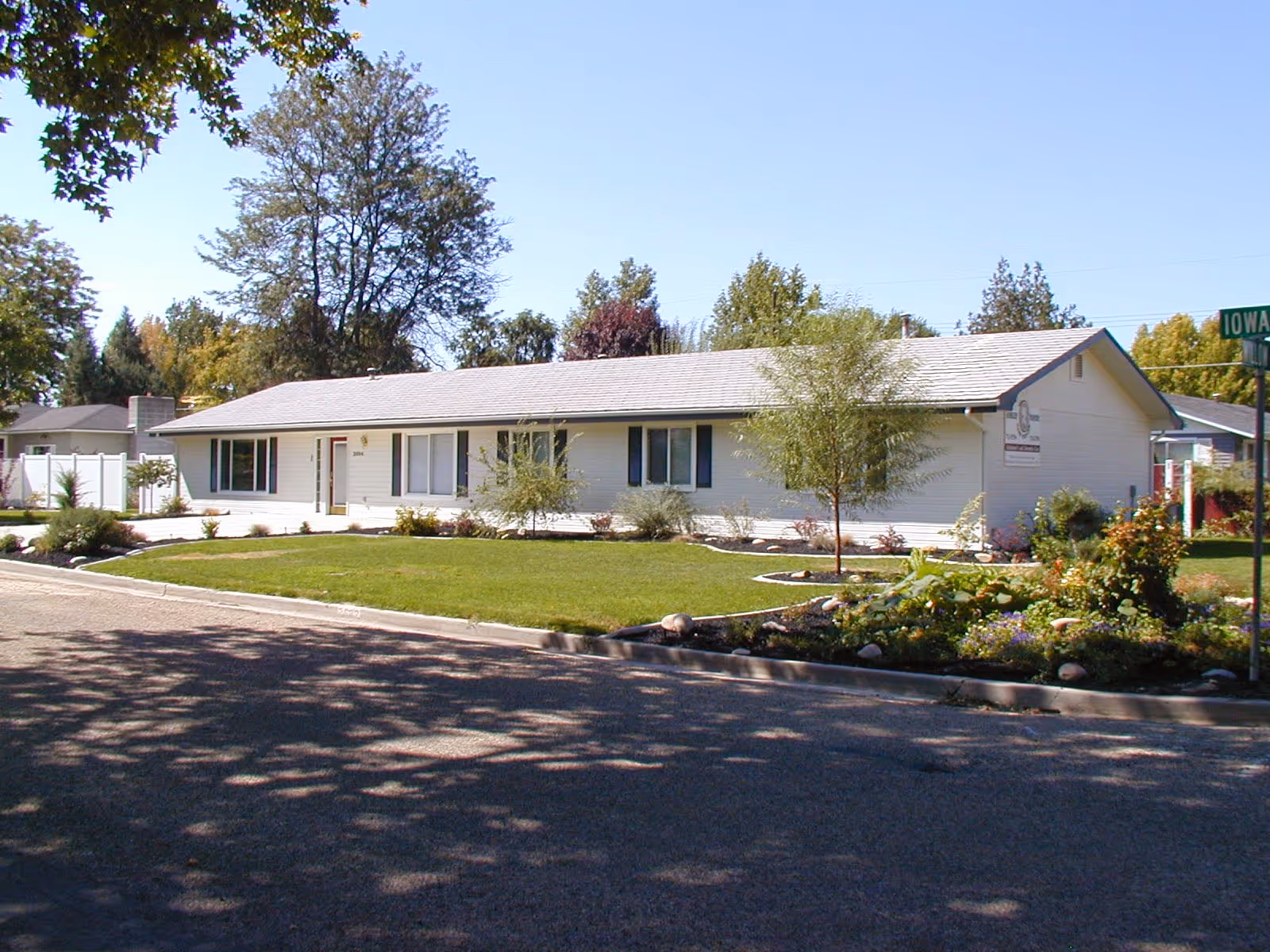 Single-story white building with a gray roof surrounded by a well-maintained lawn and landscaped garden beds, located on a corner lot with a street sign reading Iowa St.