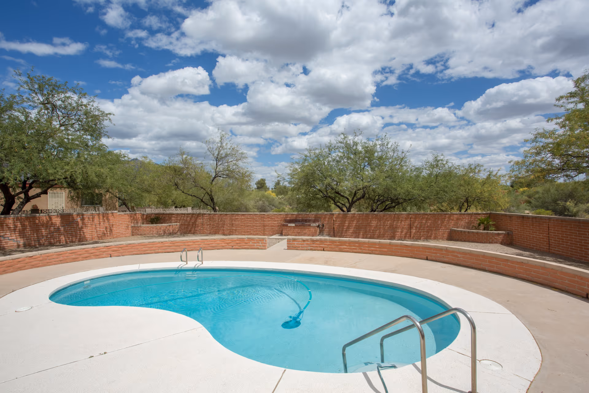 Outdoor swimming pool with clear blue water surrounded by a curved brick wall and desert trees under a partly cloudy sky.