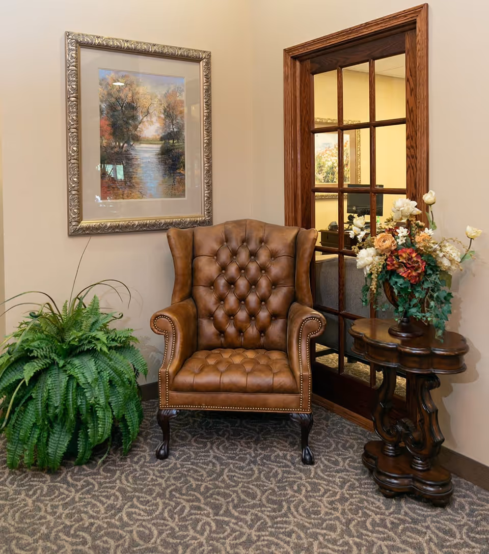 A cozy corner with a brown tufted leather armchair placed on a patterned carpet. To the left of the chair is a green fern plant, and to the right is a small wooden table with a floral arrangement. Behind the chair is a wooden-framed glass door and a framed painting of a serene river scene with trees.