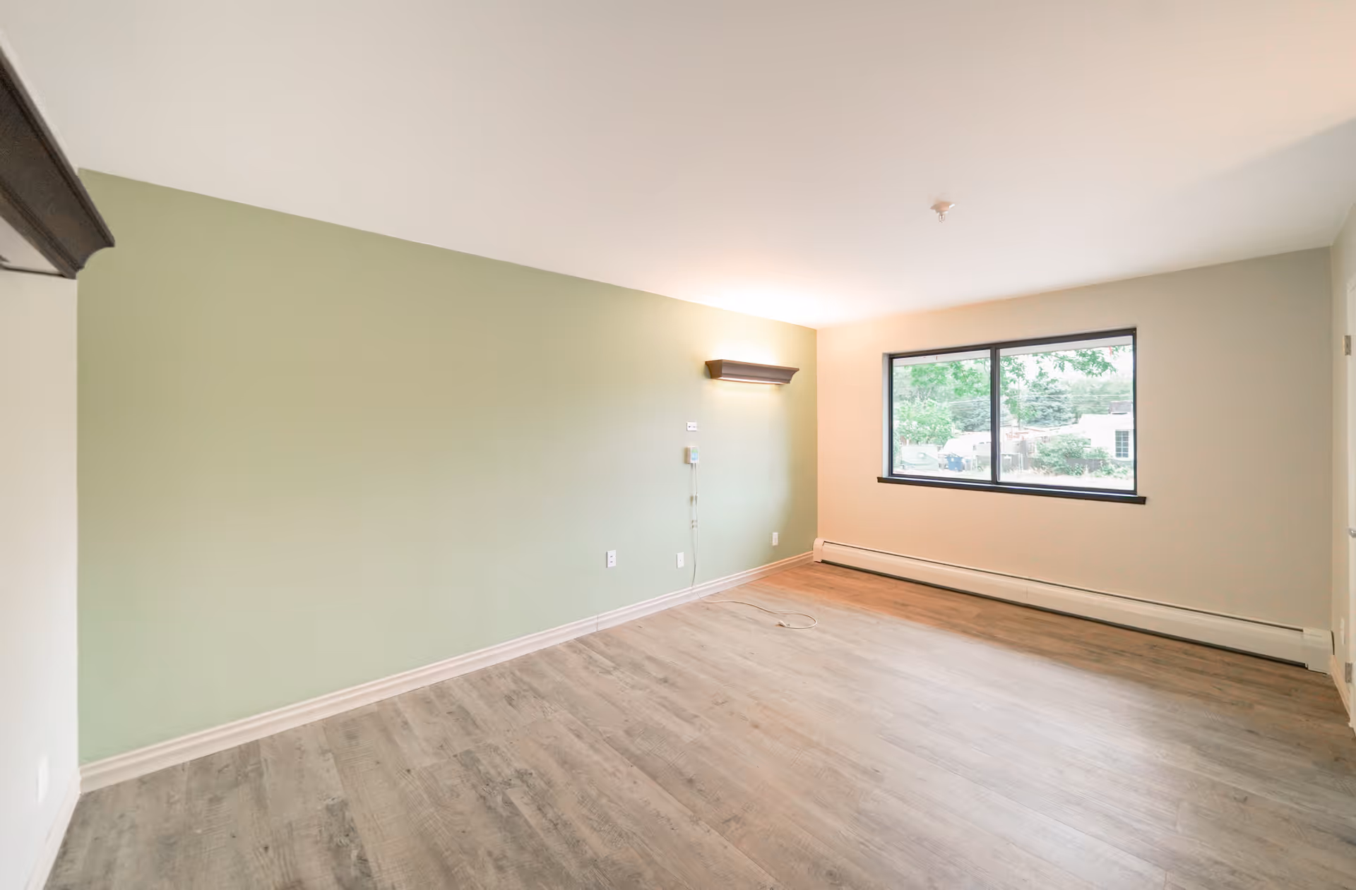 Empty room with light green and beige walls, a large window showing greenery outside, wood-like flooring, and a wall-mounted light fixture.