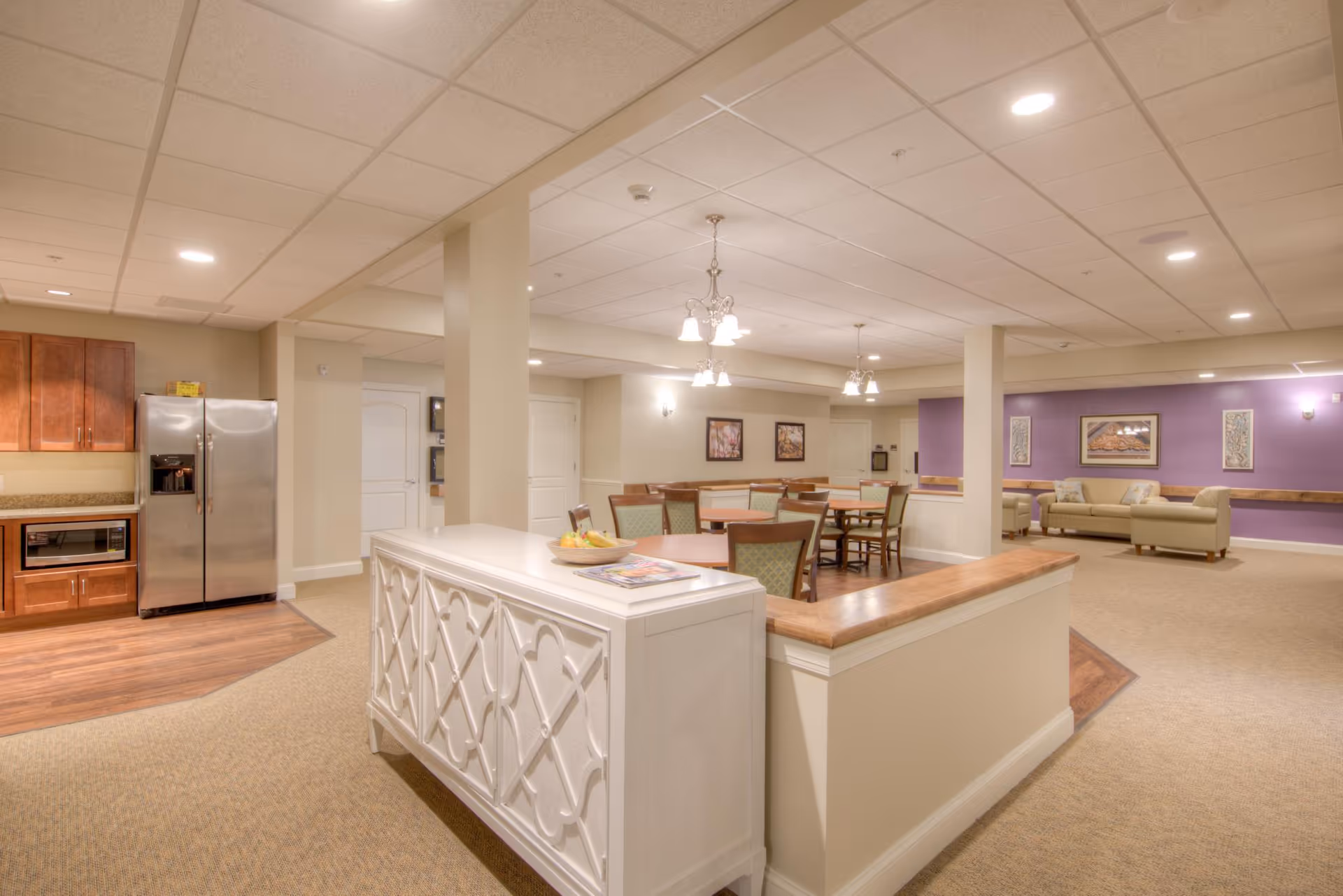 Interior view of a senior living facility showing a spacious common area with a kitchen section featuring a stainless steel refrigerator and microwave, a dining area with tables and chairs, and a lounge area with sofas against a purple accent wall. The space is well-lit with ceiling lights and chandeliers, and decorated with framed artwork on the walls.