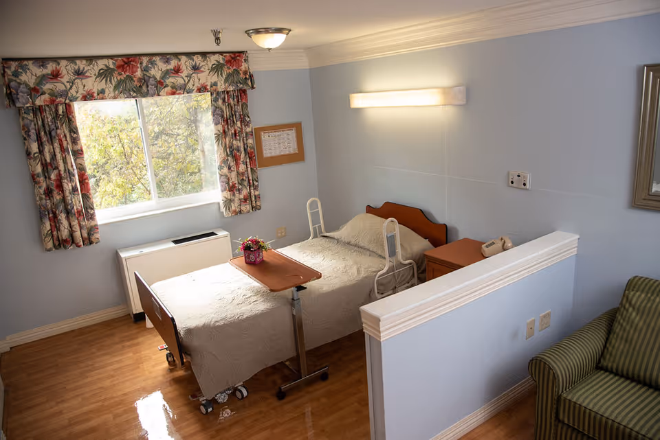 A small, clean bedroom in a senior living facility with a single hospital-style bed covered with a beige quilt. The bed has side rails and an overbed table with a small flower pot on it. There is a window with floral curtains letting in natural light, a nightstand with a telephone, a wall-mounted light above the bed, and a green striped armchair partially visible behind a half wall.