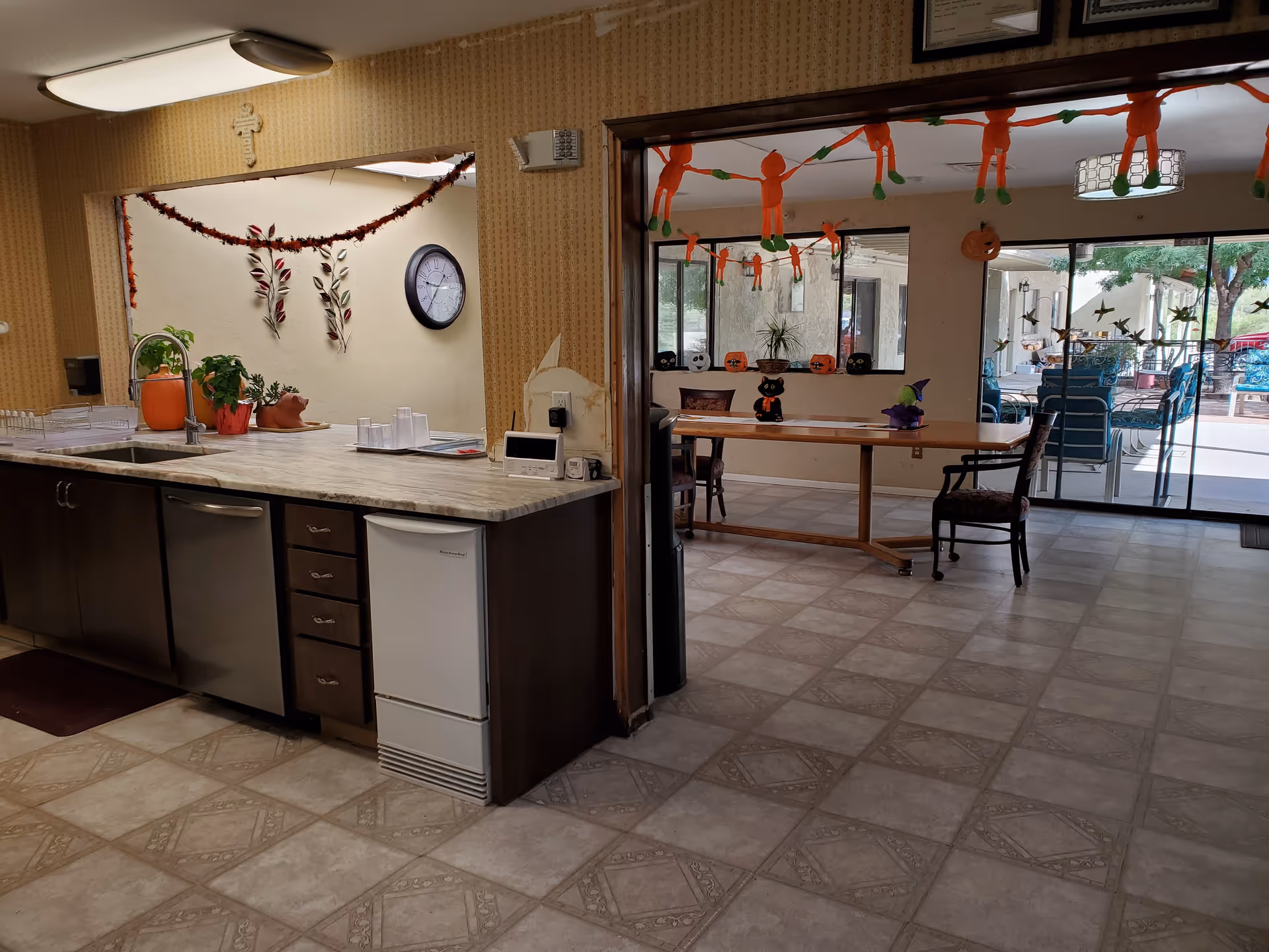Interior view of a senior living common area with a kitchenette counter, a long table, Halloween decorations, and sliding glass doors to an outdoor patio.
