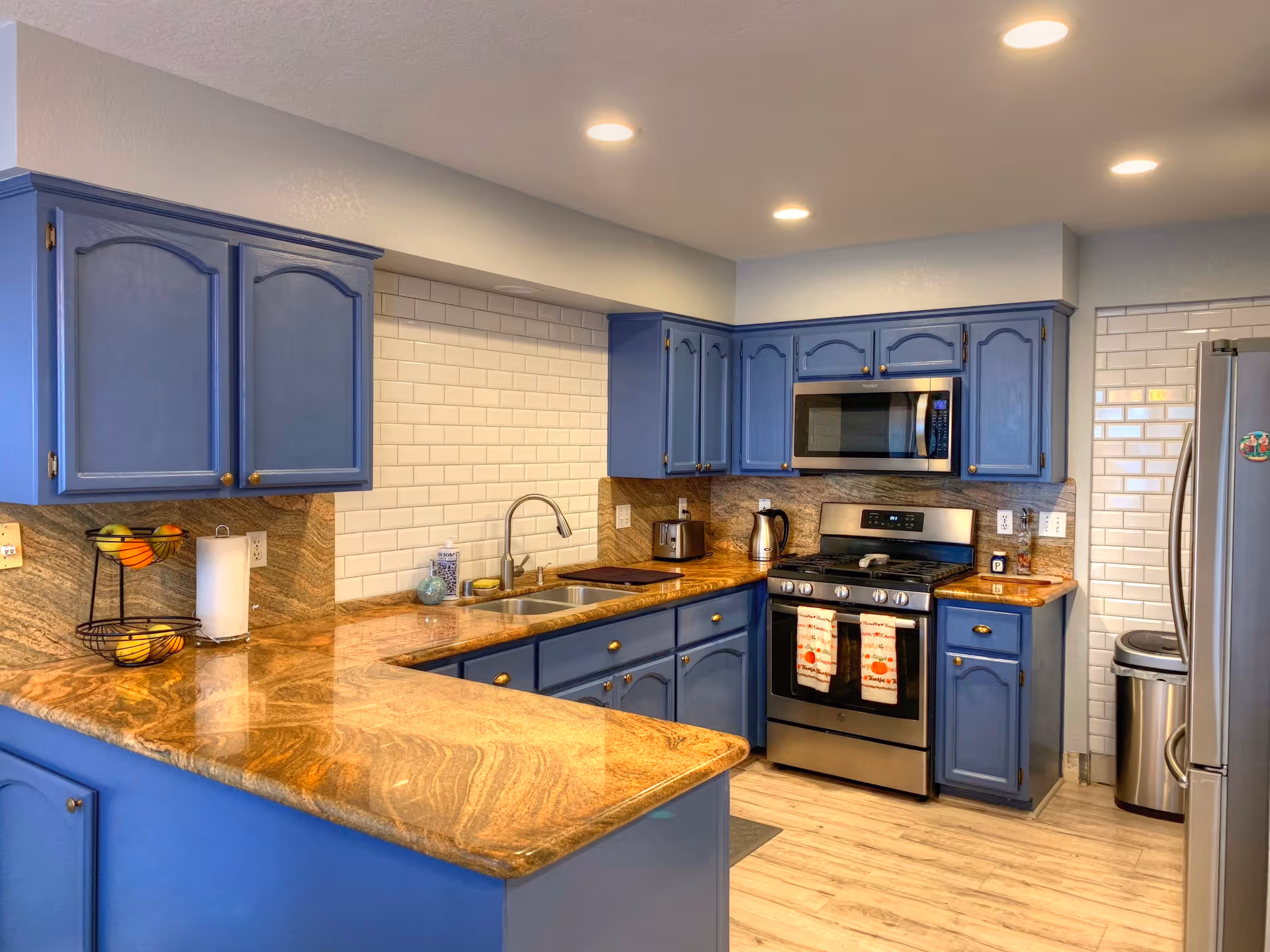 A modern kitchen with blue cabinets, granite countertops, and stainless steel appliances including a stove, microwave, and refrigerator. The backsplash features white subway tiles, and the kitchen is well-lit with recessed ceiling lights. There is a double sink, a fruit basket, a paper towel holder, and small kitchen appliances on the counter.