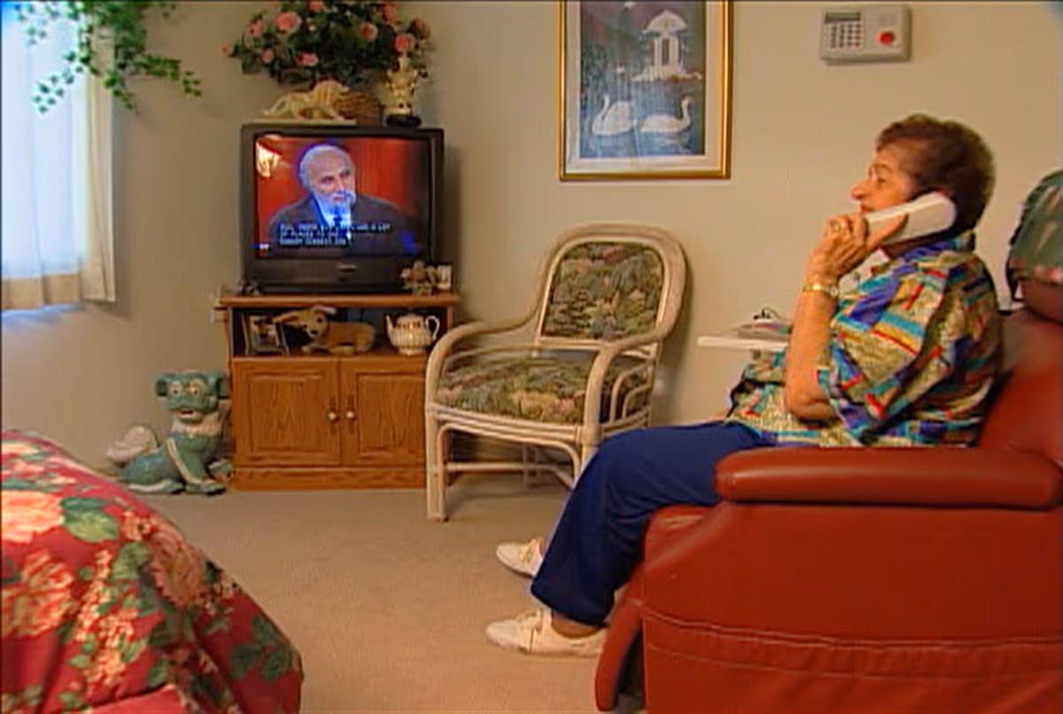 An elderly woman sits on a red recliner talking on the phone in a cozy room with a TV, wicker chair, and floral bedspread.