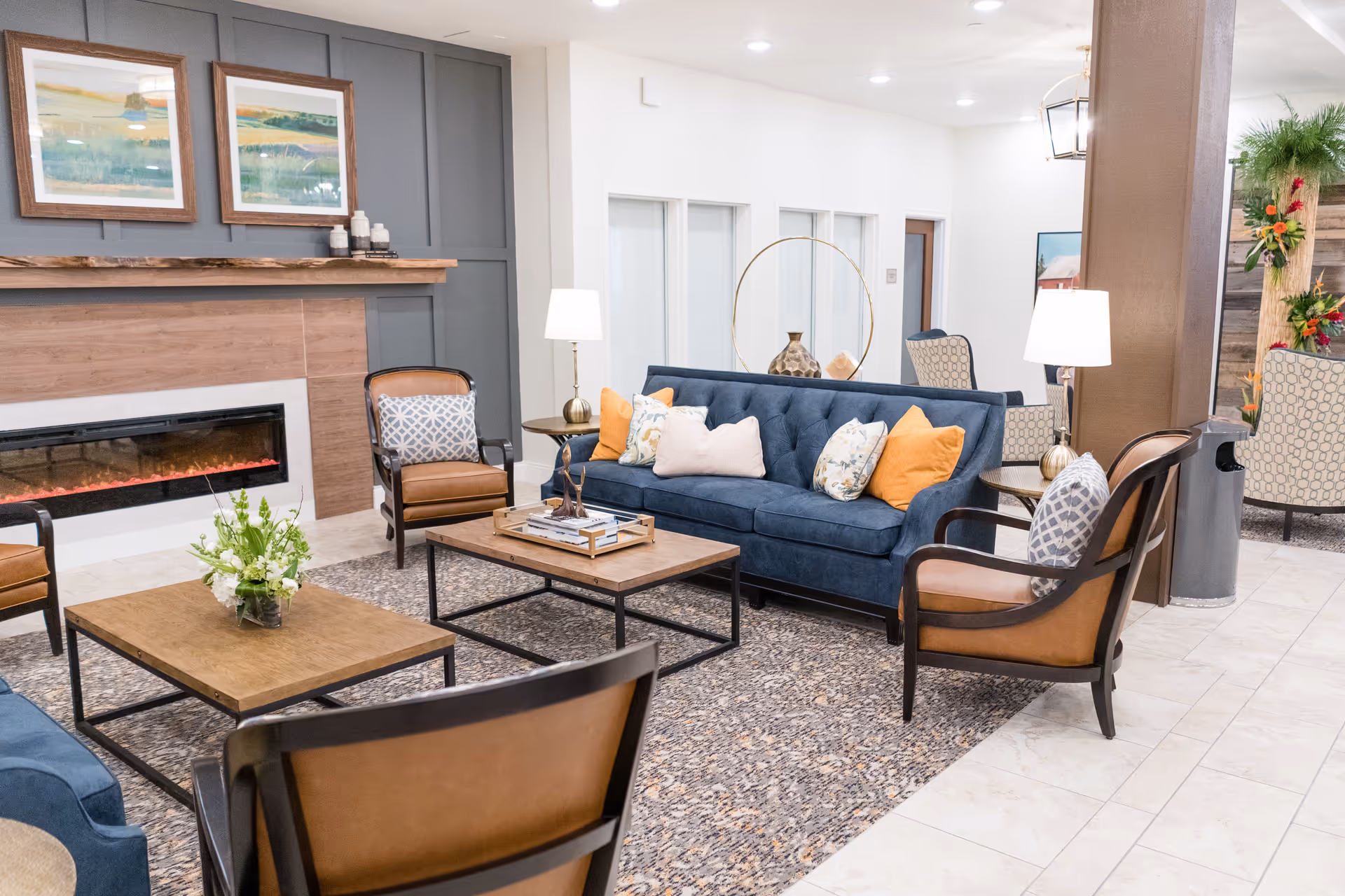 A cozy living room area with a blue sofa adorned with colorful pillows, surrounded by brown leather chairs with patterned cushions. There are two wooden coffee tables, one with a vase of flowers and the other with decorative items. A modern electric fireplace is set into a wooden and gray paneled wall, above which hang two landscape paintings. The room is well-lit with table lamps and ceiling lights, and there is a patterned rug on the tiled floor.