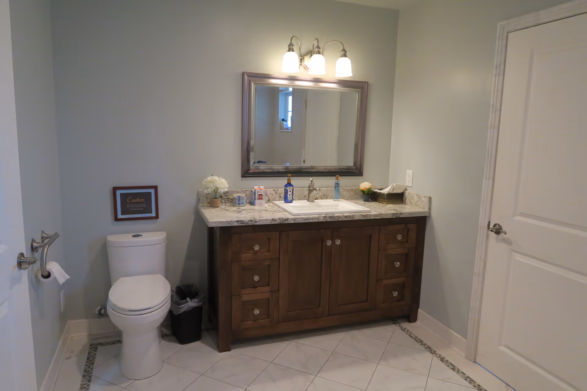 A clean and modern bathroom featuring a white toilet with a toilet paper holder on the left wall, a wooden vanity with a granite countertop, a white sink, and a large rectangular mirror above it. The vanity has multiple drawers and cabinets with crystal knobs. There are decorative items on the countertop including flowers, soap dispensers, and a tissue box. The floor is tiled with white tiles bordered by a decorative pattern. A closed white door is visible on the right side of the image.