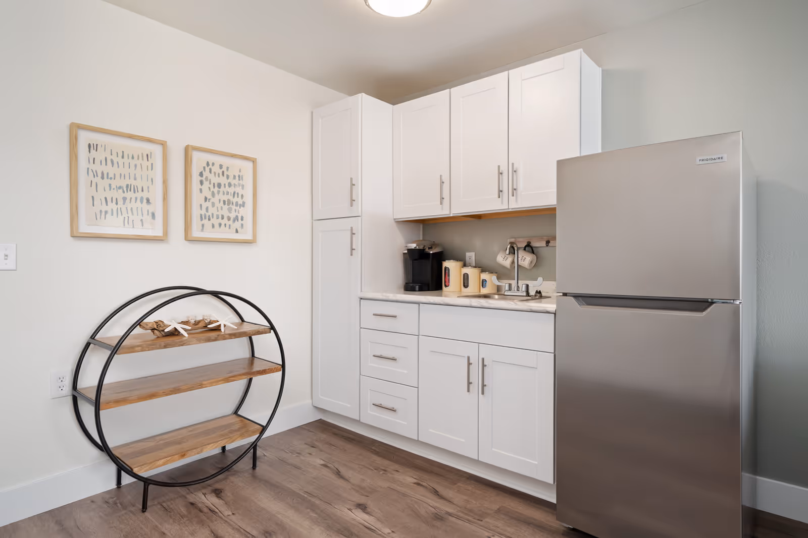A small kitchen area with white cabinets, a stainless steel refrigerator, a coffee maker, and a small sink. There is a round wooden shelf unit with three shelves on the left side and two framed abstract art pieces hanging on the wall above it. The floor is wood with a light finish.