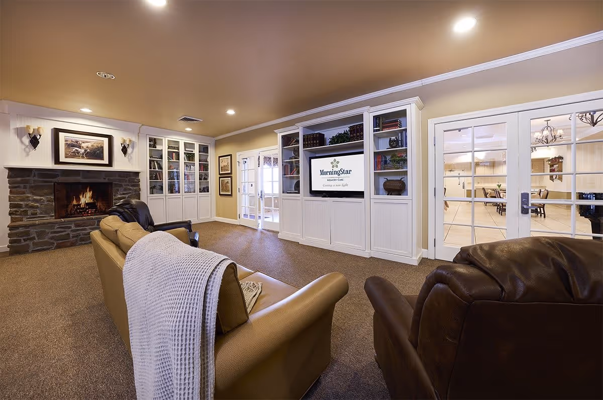 A cozy living room area in a senior living facility with a stone fireplace on the left, a tan sofa with a white throw blanket, and a dark brown leather armchair. Built-in white shelves and cabinets surround a TV displaying the MorningStar Memory Care logo. French doors lead to another room with tables and chairs.