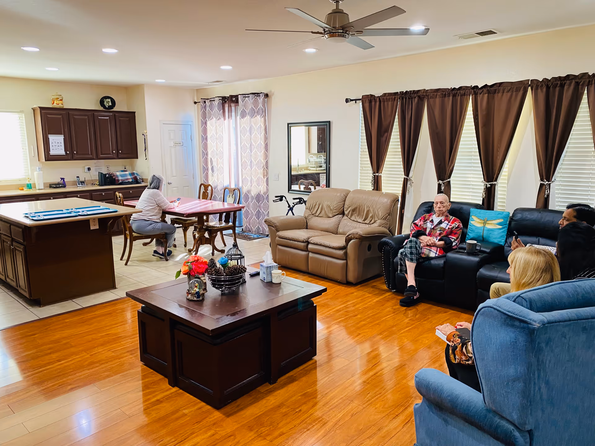Communal living room and kitchen area with sofas, a coffee table, a dining table, and several people seated.
