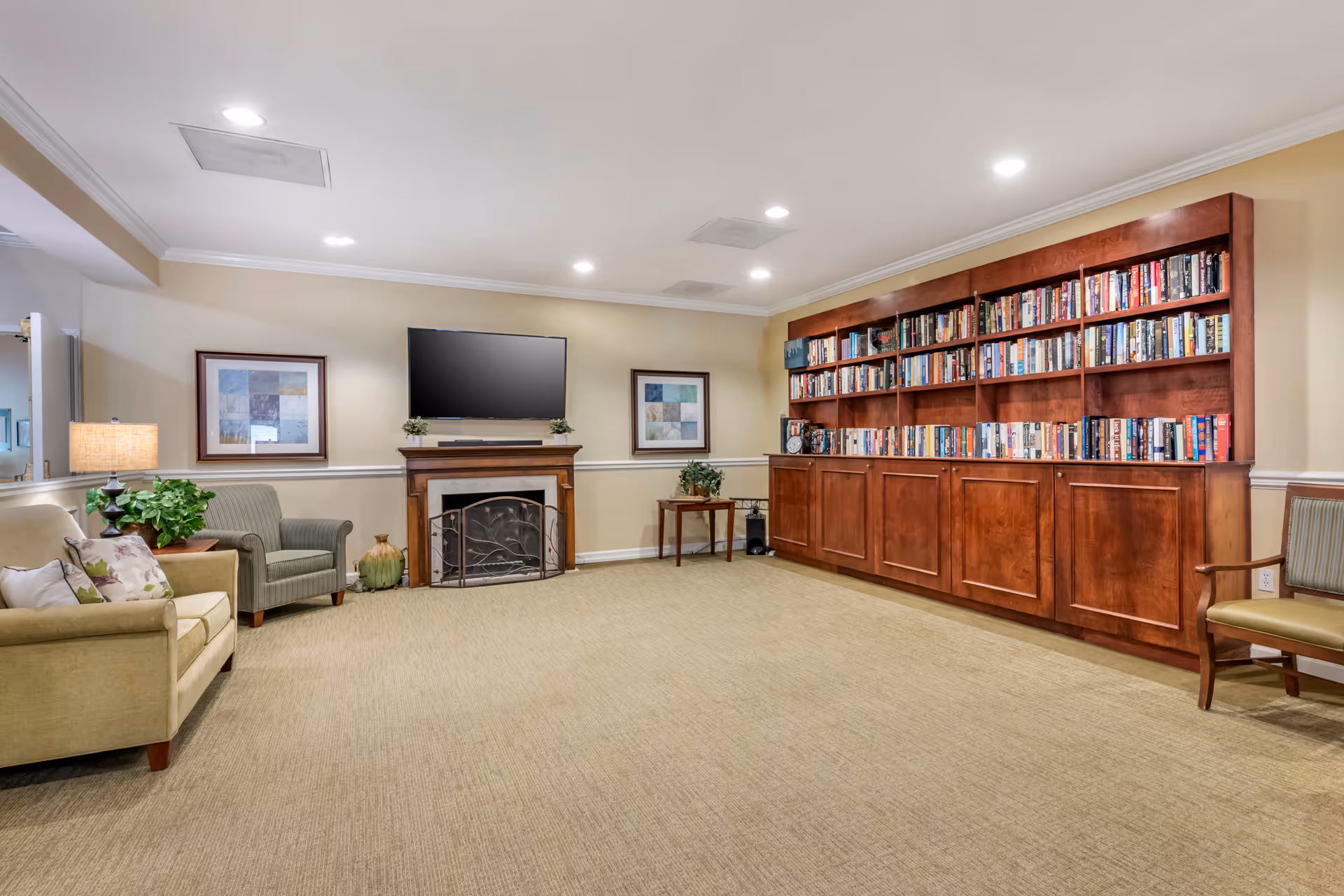 A cozy living room area with beige carpet and cream-colored walls. The room features a wooden fireplace with a flat-screen TV mounted above it, flanked by two framed artworks. To the left, there is a beige sofa with floral cushions and a green armchair next to a side table with a lamp and a plant. On the right side, there is a large wooden bookshelf filled with books and a wooden chair with a green cushion. The room is well-lit with recessed ceiling lights.