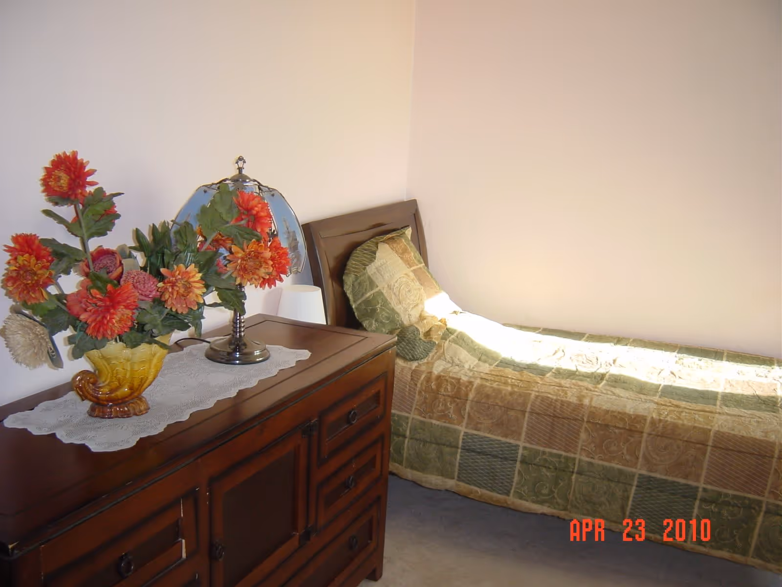 A small bedroom with a single bed covered with a patchwork quilt in shades of green and beige. Next to the bed is a wooden dresser with a lace runner, a decorative vase with red and orange flowers, and a vintage-style lamp with a stained glass shade. The walls are plain and light-colored.