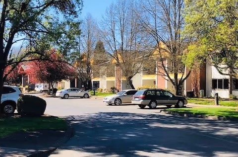 Exterior view of a senior living facility with a parking lot in front. Several cars are parked along the driveway, and the building is surrounded by trees with some leaves turning colors, indicating early spring or fall.