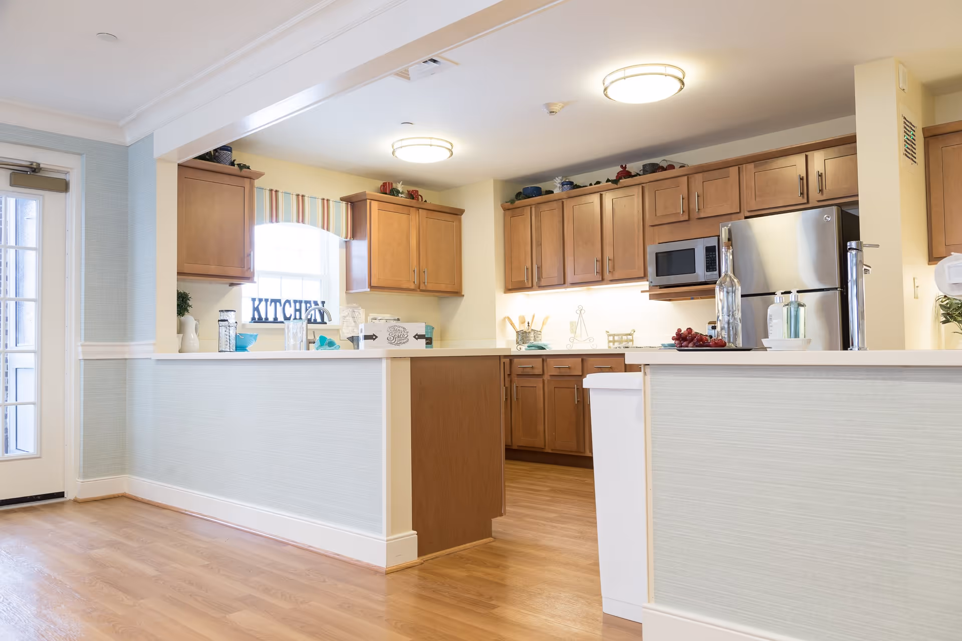 Bright and clean kitchen area with wooden cabinets, stainless steel refrigerator, microwave, and a white countertop island. There is a window with a striped valance and a decorative sign that says 'KITCHEN'. The floor is wooden, and the walls are painted light yellow and light blue. The kitchen is well-lit with ceiling lights.