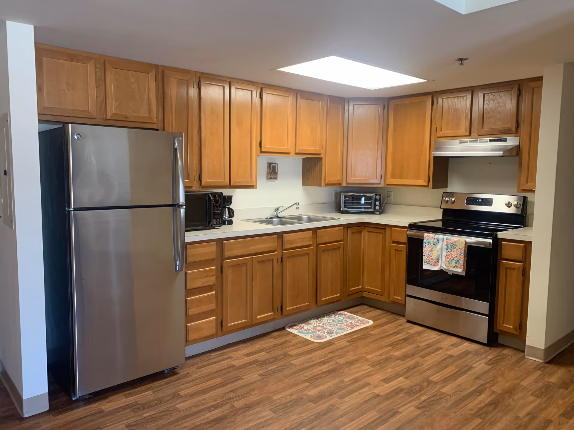 A clean kitchen with wooden cabinets, a stainless steel refrigerator, a stainless steel electric stove with two floral-patterned towels hanging on the handle, a double sink, a microwave, a coffee maker, a toaster oven, and a colorful floor mat in front of the sink. The floor is wood-patterned and the ceiling has a large rectangular light fixture.