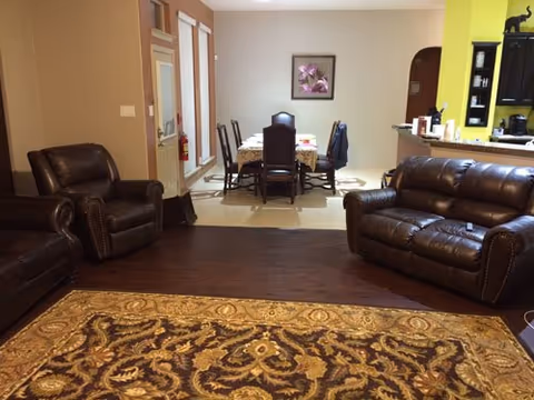 A cozy living room with dark brown leather sofas and armchairs arranged around a patterned area rug. In the background, there is a dining area with a table and chairs, a framed floral picture on the wall, and a kitchen counter with various items on it.