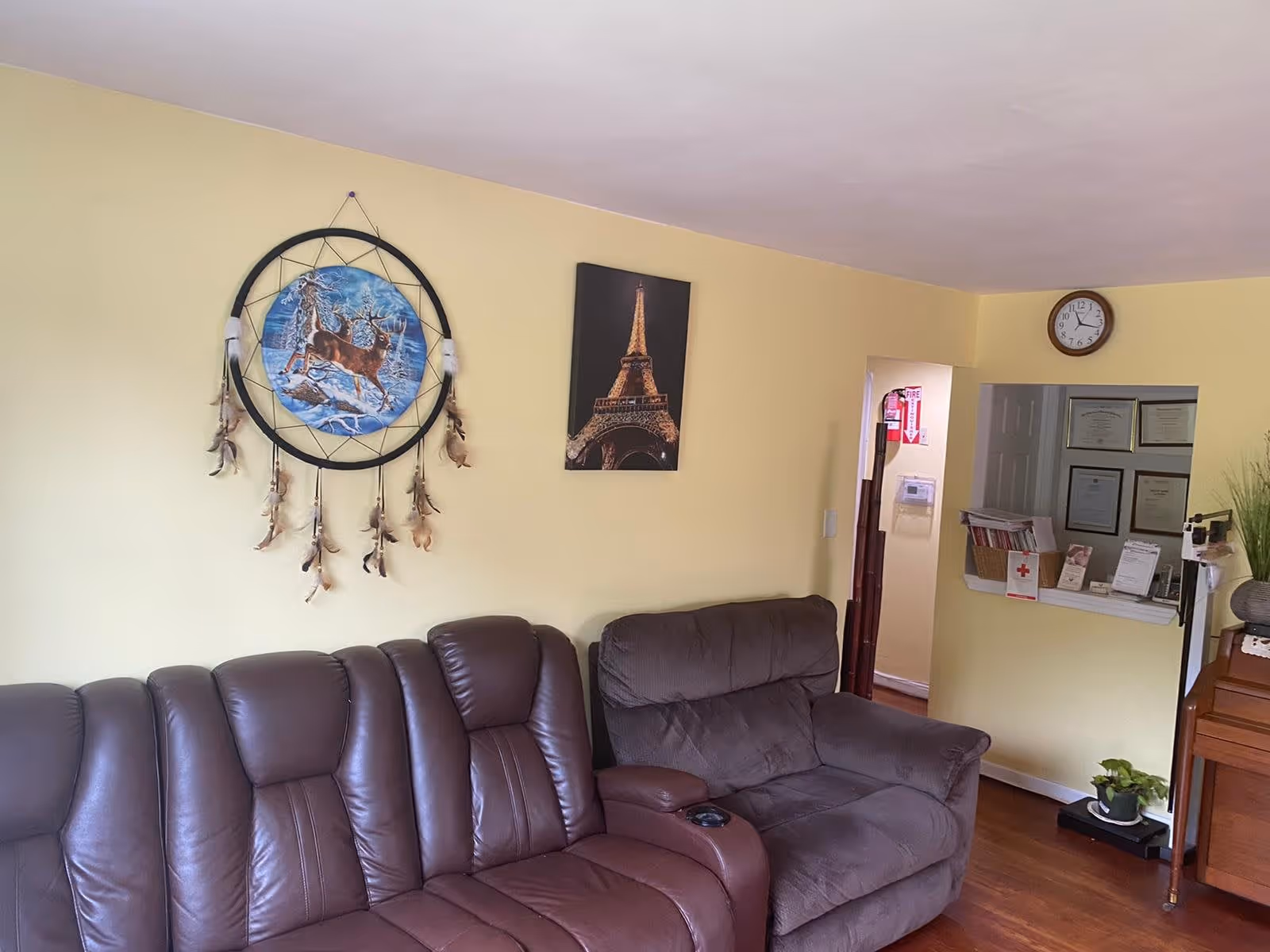 A cozy living room with two brown leather sofas against a light yellow wall. On the wall, there is a large dreamcatcher with a deer design and a framed picture of the Eiffel Tower. A clock is mounted above a small reception window with certificates displayed inside. A small plant and a piano are visible on the right side of the room.