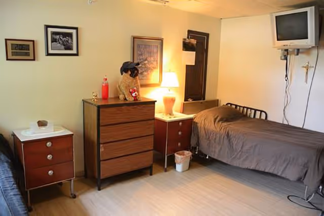 A simple bedroom with two single beds, each with brown bedding. Between the beds are two wooden nightstands and a wooden dresser with a stuffed dog wearing a hat on top. A lamp is lit on one of the nightstands. A small television is mounted on the wall above one bed. The room has light-colored walls and a wooden floor.