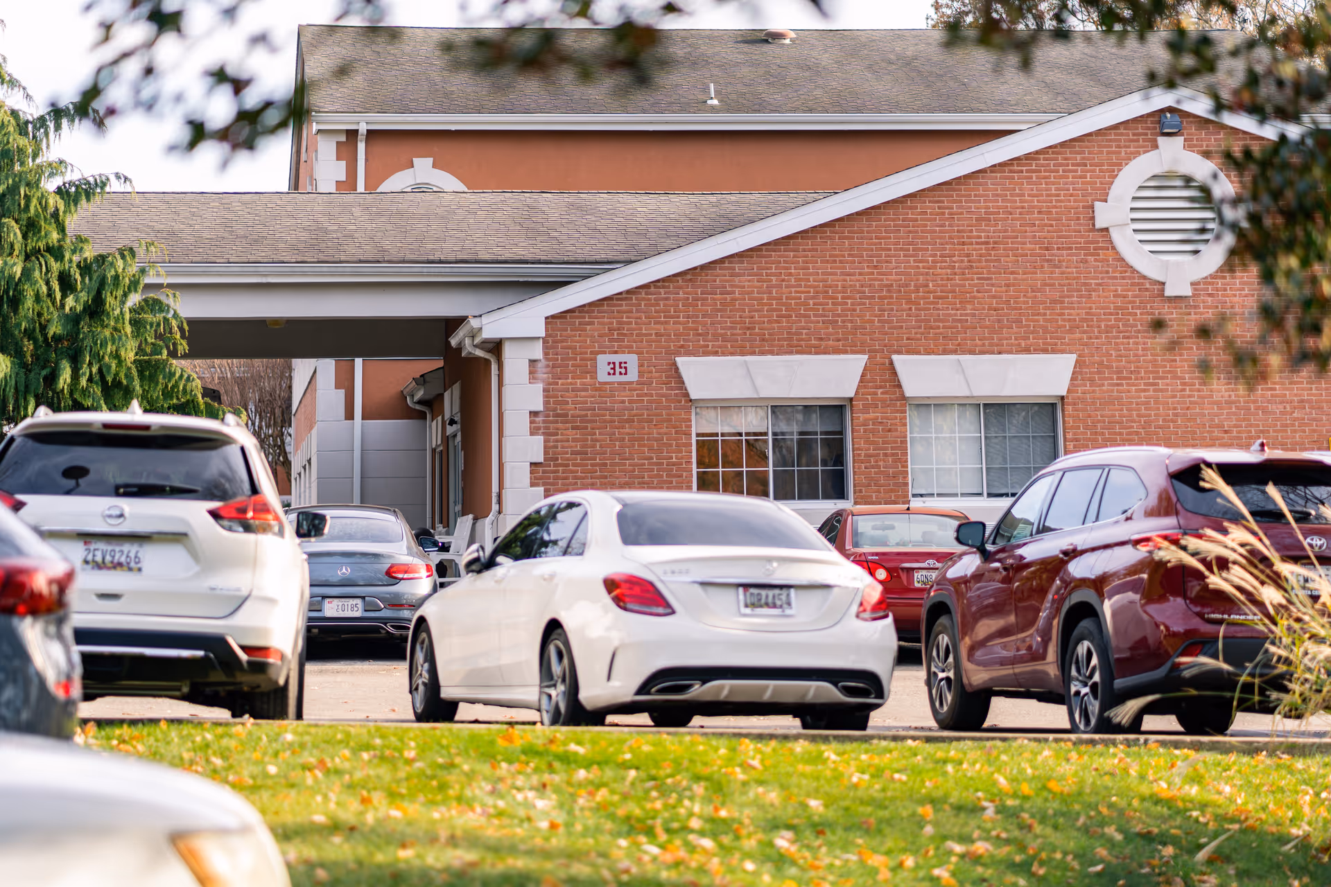 Parking lot with several cars parked in front of a brick building with white trim and windows, partially obscured by tree branches and leaves.