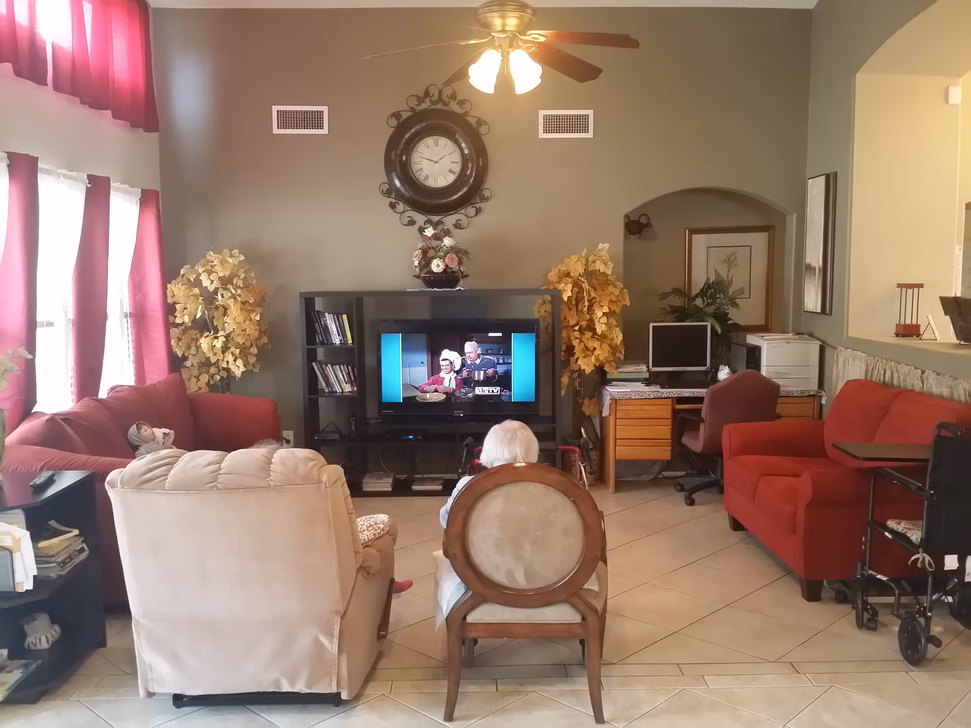 A cozy living room in a senior living facility with two elderly people seated facing a television. The room features a beige recliner, a wooden chair, a red sofa, a TV stand with a television playing a show, decorative plants, a wall clock, a ceiling fan with lights, and a desk with a computer and printer.