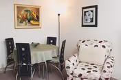 A small dining area with a round table covered by a green tablecloth and four black chairs. Next to the table is a cushioned armchair with a white cover featuring a brown and beige leaf pattern. The walls are white and decorated with two framed pictures, one depicting fruit and the other a black and white floral image. A tall floor lamp stands in the corner providing light.