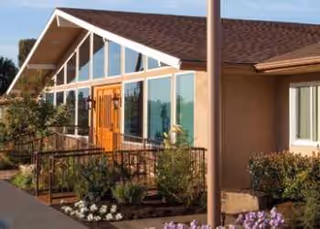 Sunlit exterior of a modern senior care building with glass walls and wooden doors.