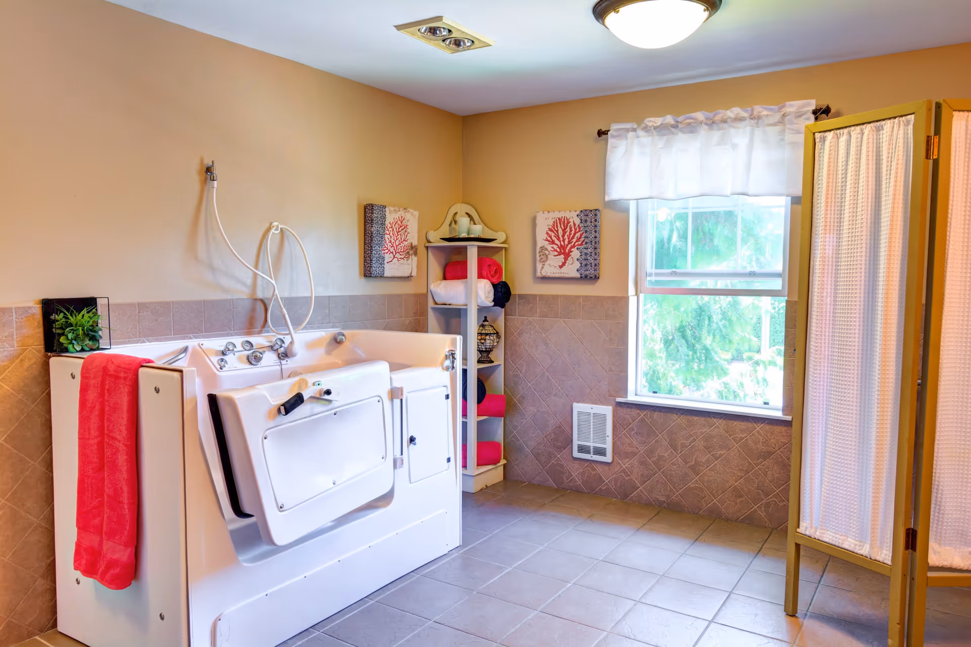 A spacious bathroom featuring a walk-in bathtub with a red towel hanging on the side. There is a white shelving unit in the corner holding rolled towels and decorative items. The room has beige walls with tiled lower half, a window with a white curtain, and a folding privacy screen on the right side.