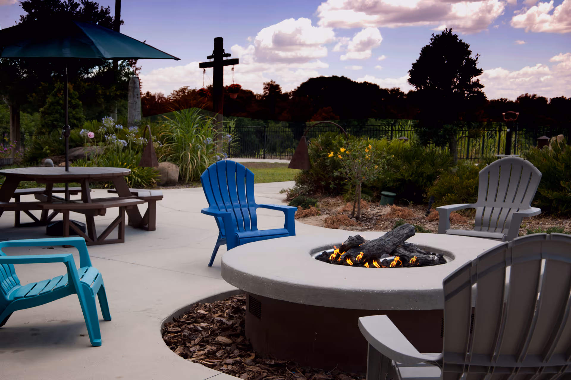 Outdoor patio area with a round fire pit surrounded by colorful Adirondack chairs in blue, teal, and gray. There is a wooden picnic table with a large green umbrella on the left side, and various plants and trees in the background under a partly cloudy sky.