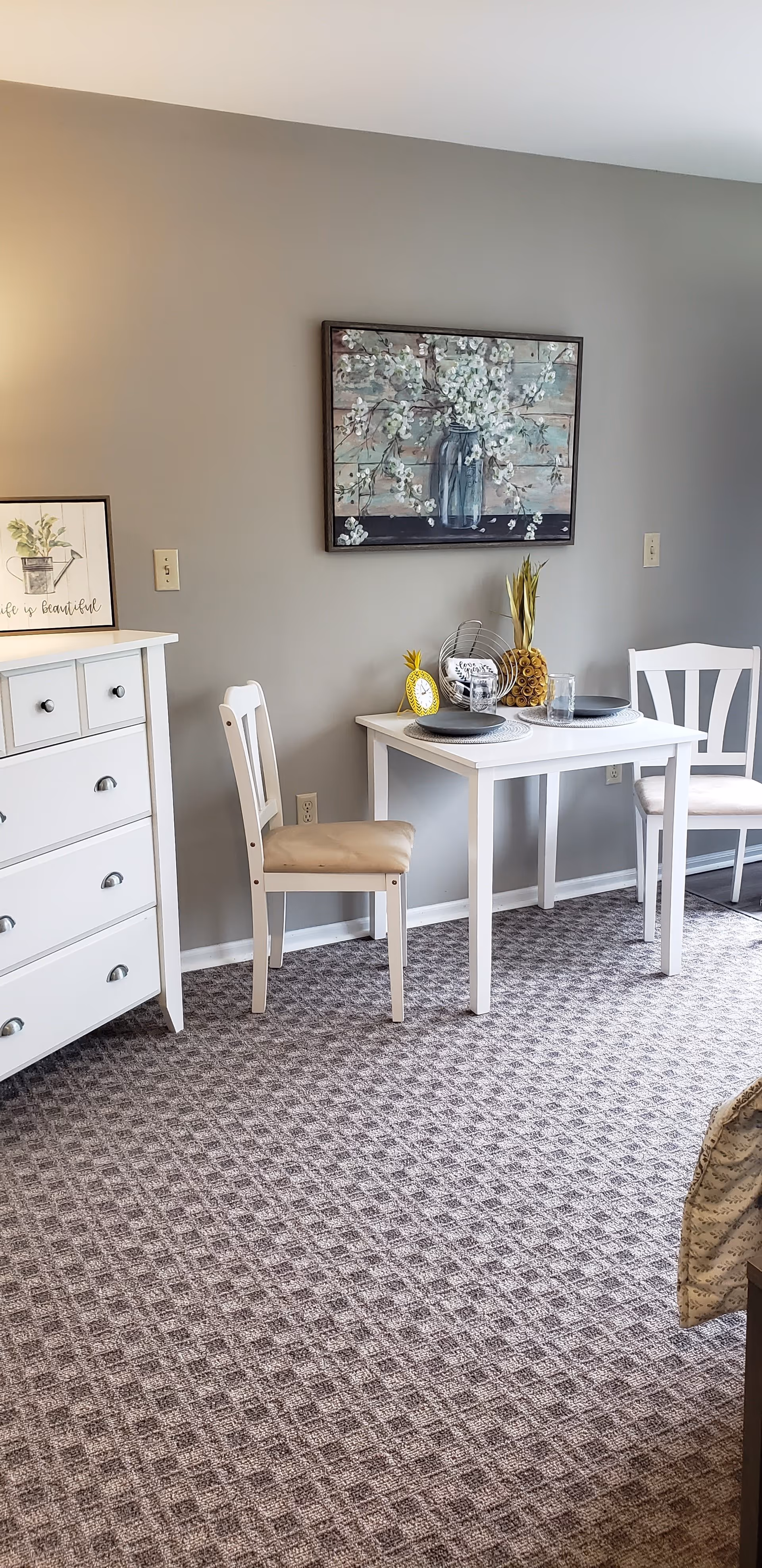 A small dining area with a white table set for two, featuring two white chairs with beige cushions. On the table are two gray plates on woven placemats, a decorative yellow pineapple clock, a wire basket with a rolled towel, and a glass. Behind the table is a wall with a painting of white flowers in a blue vase. To the left is a white dresser with drawers and a framed picture that says 'Life is Beautiful'. The floor is carpeted with a patterned design.