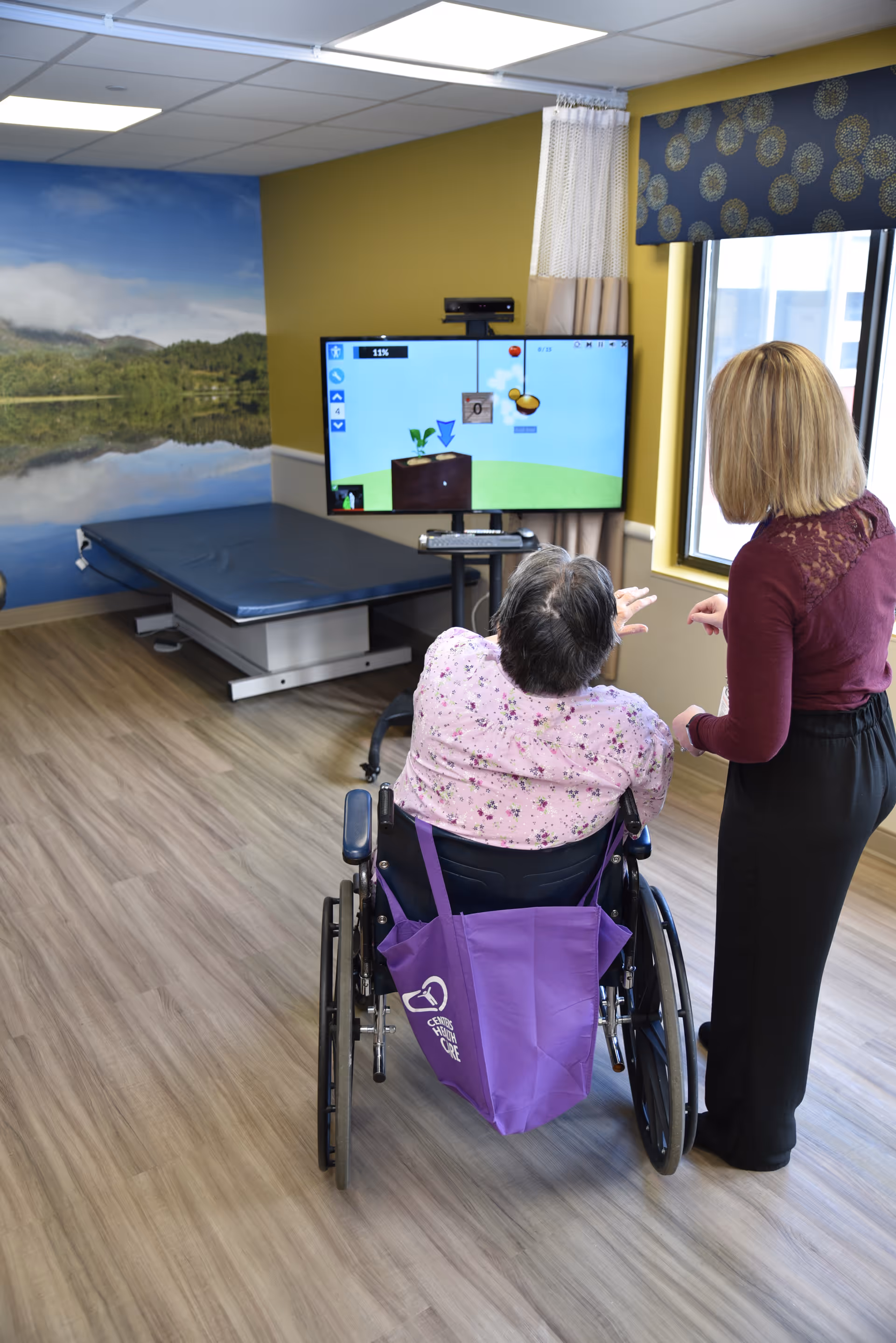 A person in a wheelchair playing an interactive game on a large screen with the assistance of a standing caregiver in a room with wood flooring, a treatment table, and a wall mural of a lake and forest.