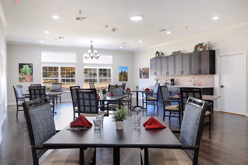 A bright dining room with multiple tables and chairs arranged neatly. Each table has a small green plant, glasses, and red folded napkins. The room features large windows letting in natural light, a chandelier, and a kitchenette area with dark cabinets and a coffee maker. Artwork is displayed on the walls.